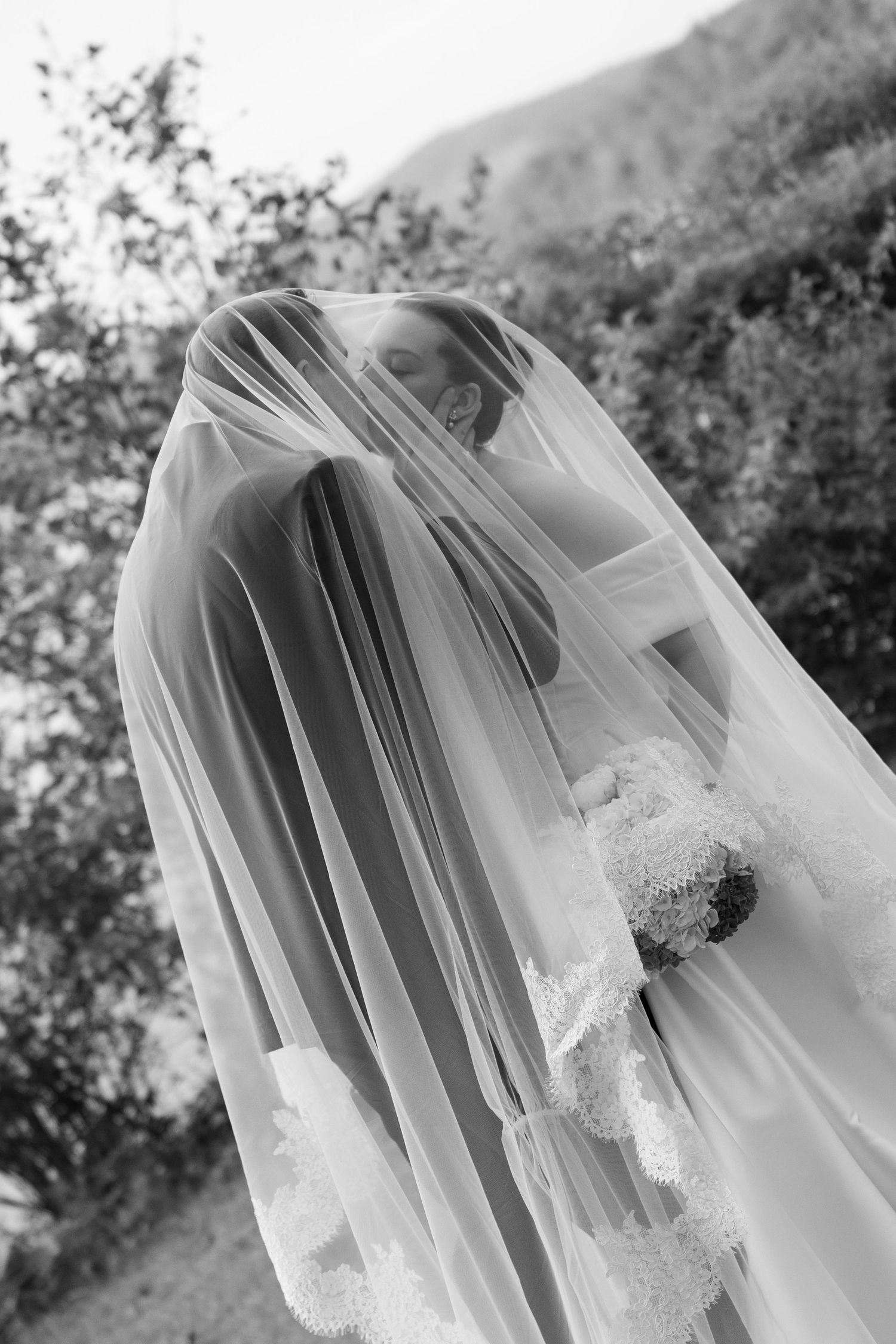 Black and white photo of a bride covered by her veil, holding a bouquet, with trees in the background.