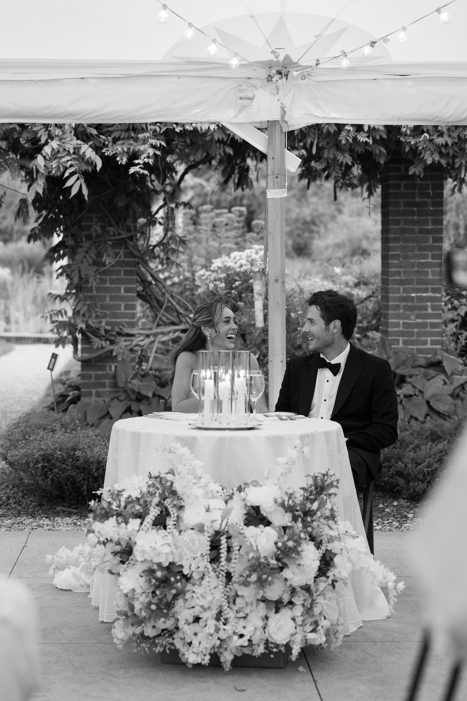 Black and white photo of a smiling bride and groom seated at a floral-adorned sweetheart table at an outdoor wedding reception.