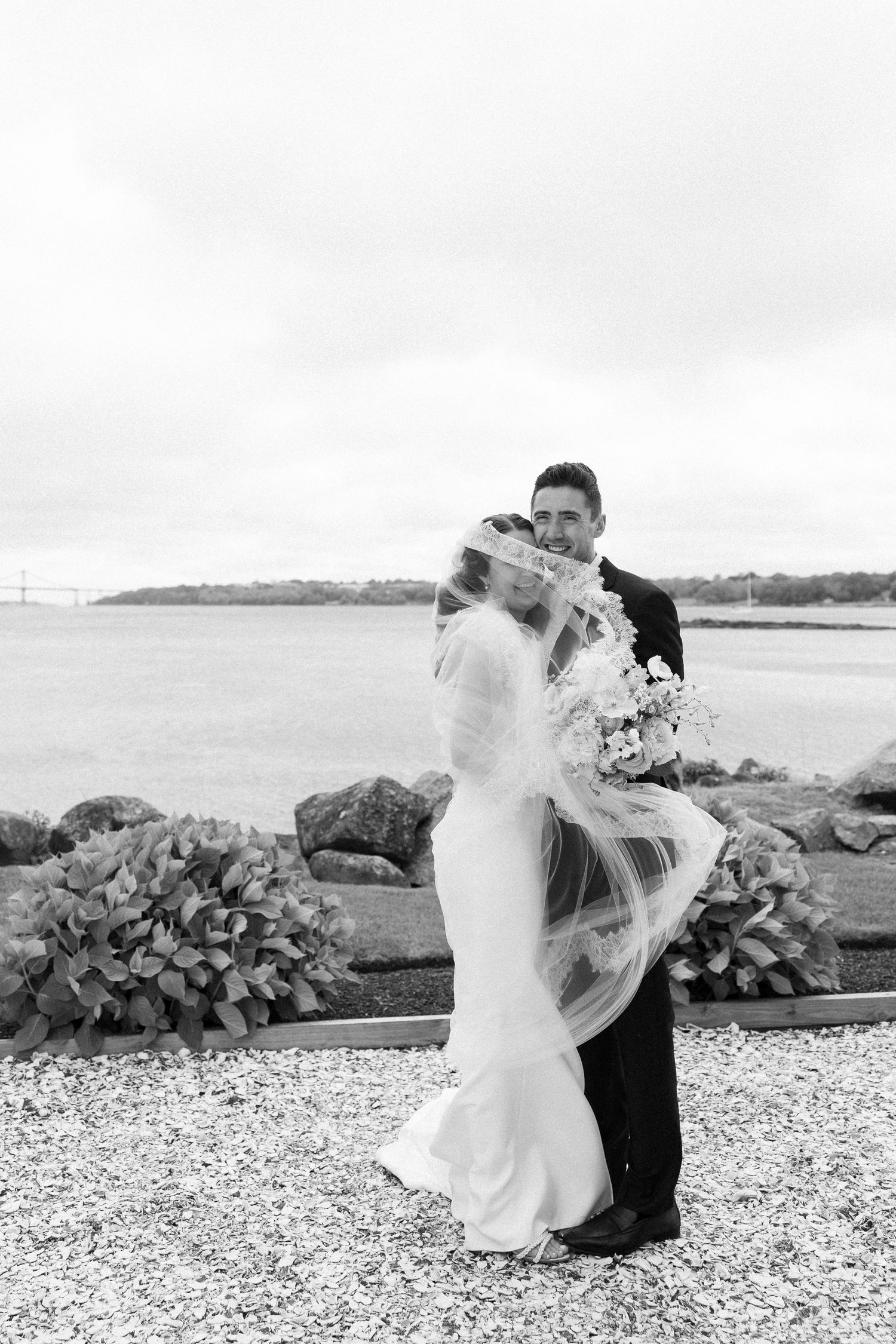 Black and white wedding portrait of bride and groom embracing by rocky shoreline with veil blowing in wind.
