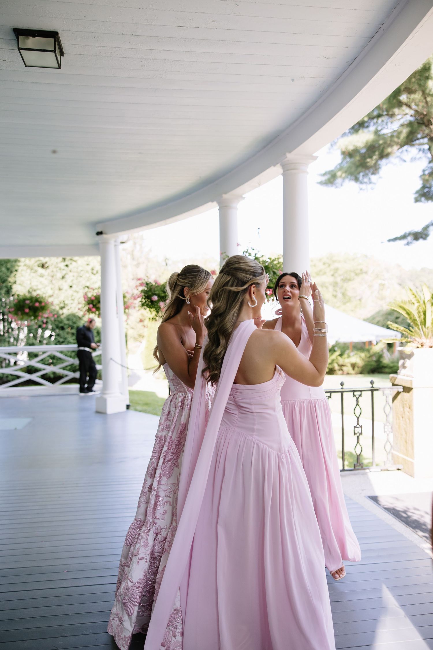 Bridesmaids in pink gowns chat on a white columned porch at a wedding venue.