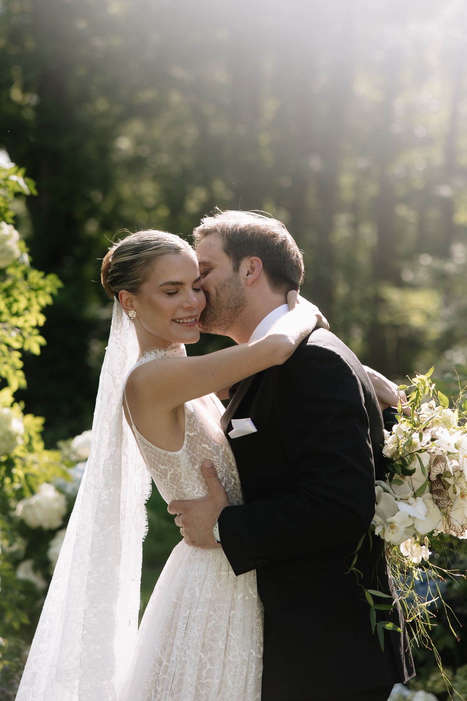 Bride and groom embracing in sunlit garden, bride wearing lace wedding dress with veil holding white floral bouquet.