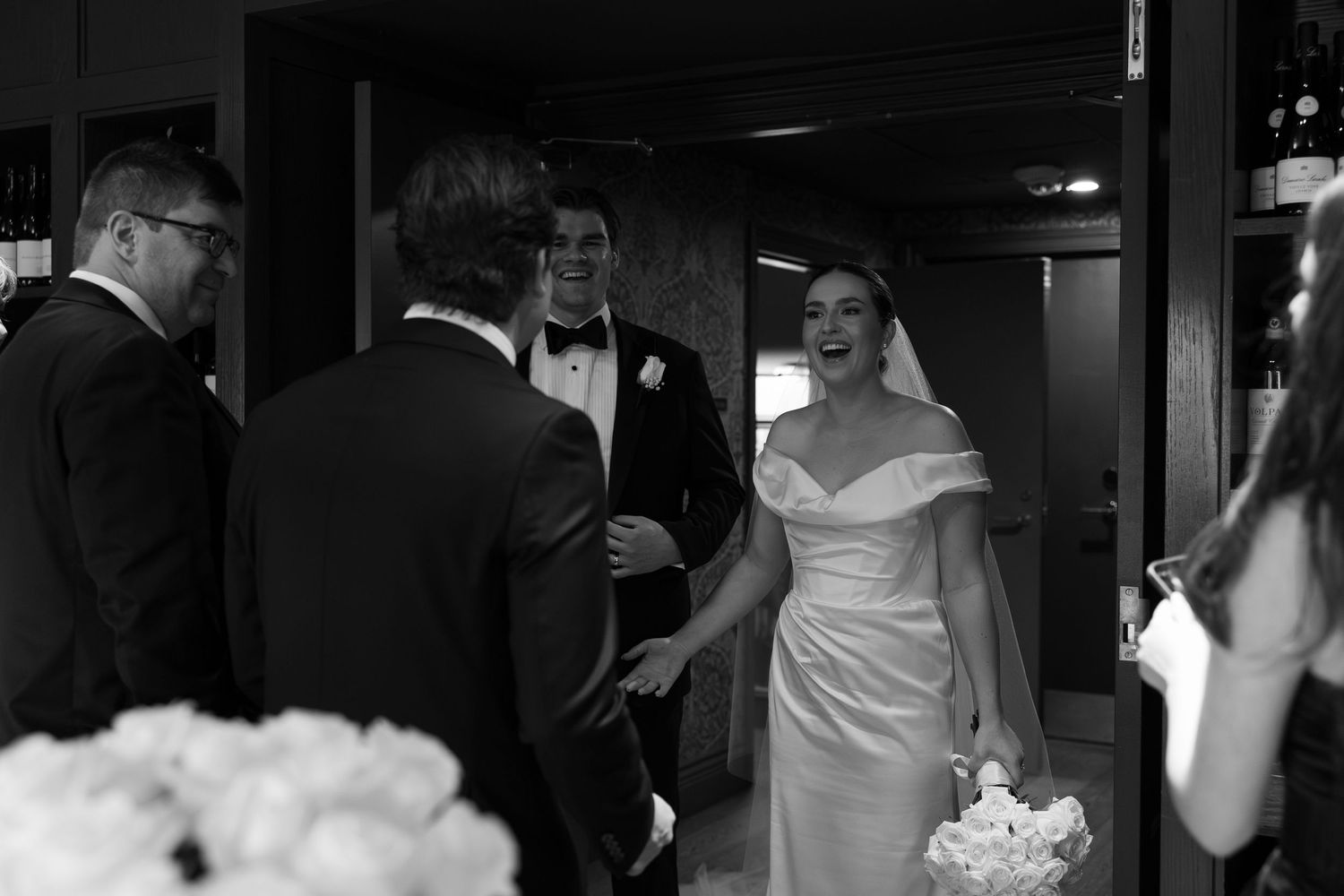 Bride in off-shoulder gown laughing with groom and guests at elegant black and white wedding reception.