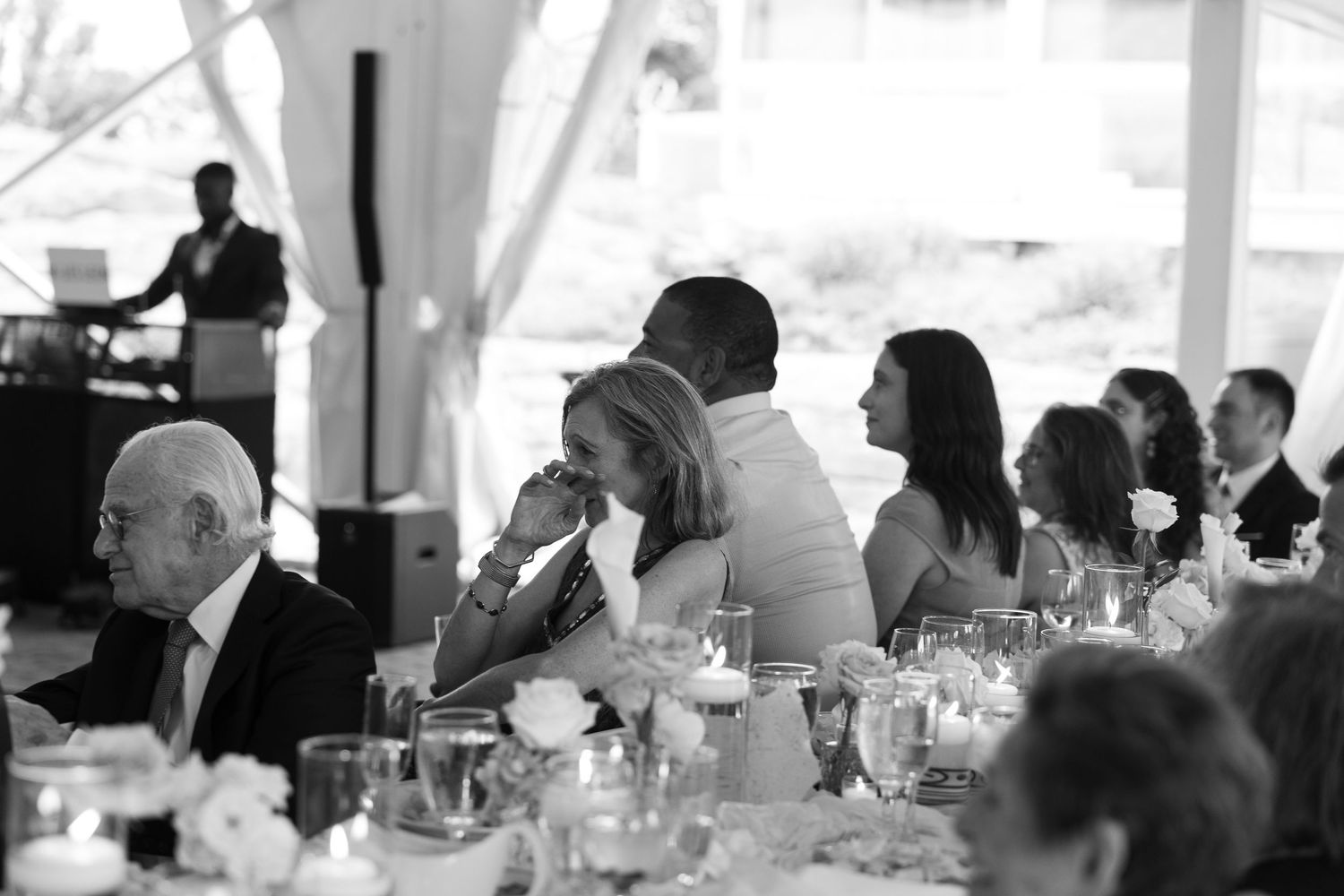 Black and white photo of wedding guests dining at reception tables under white tent.