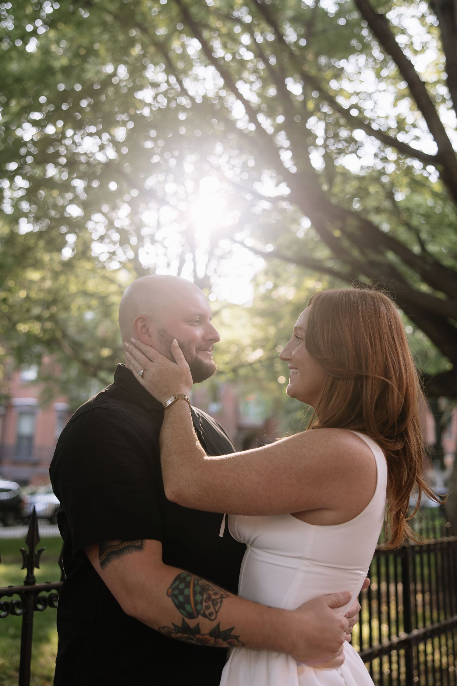 Couple sharing tender moment in sunlit park with brick buildings in background.