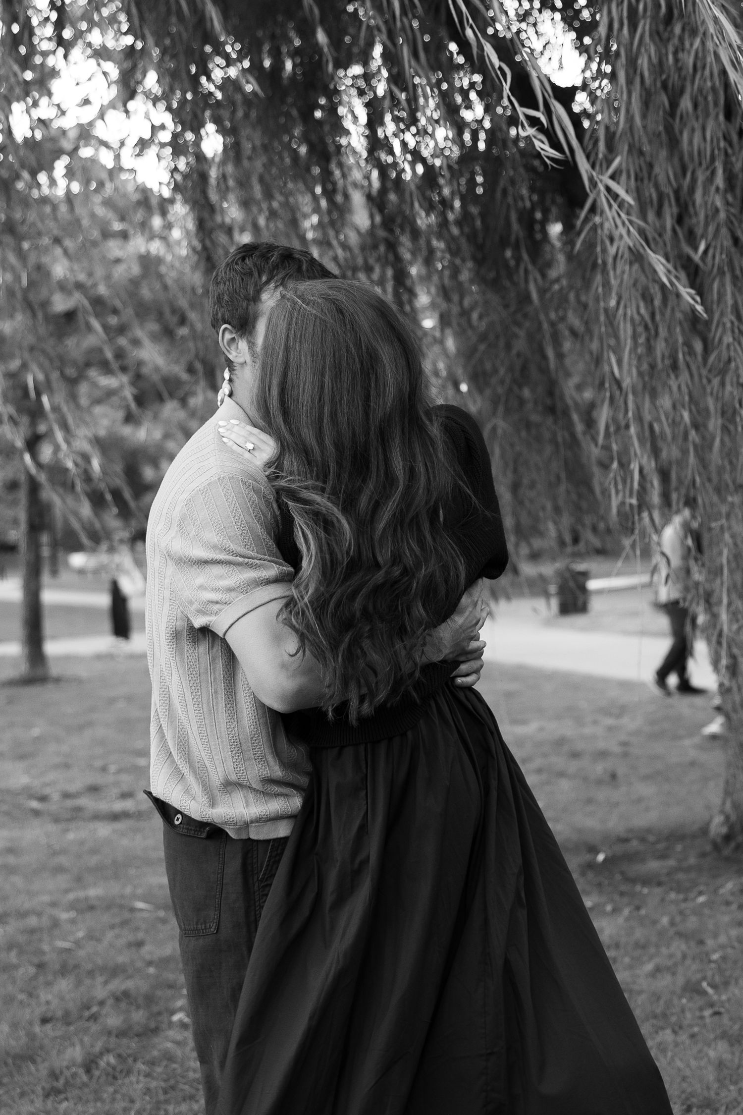 A couple sits together under a willow tree in a park, creating a romantic black and white moment.