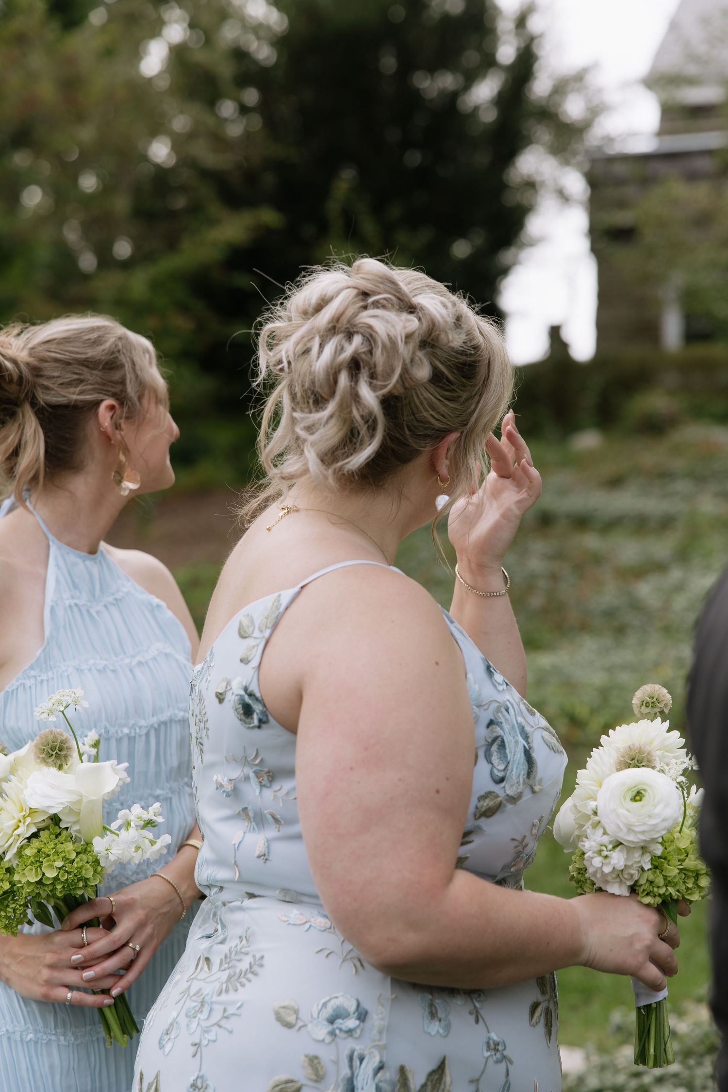 Bridesmaids in light blue floral dresses hold white bouquets during outdoor wedding.