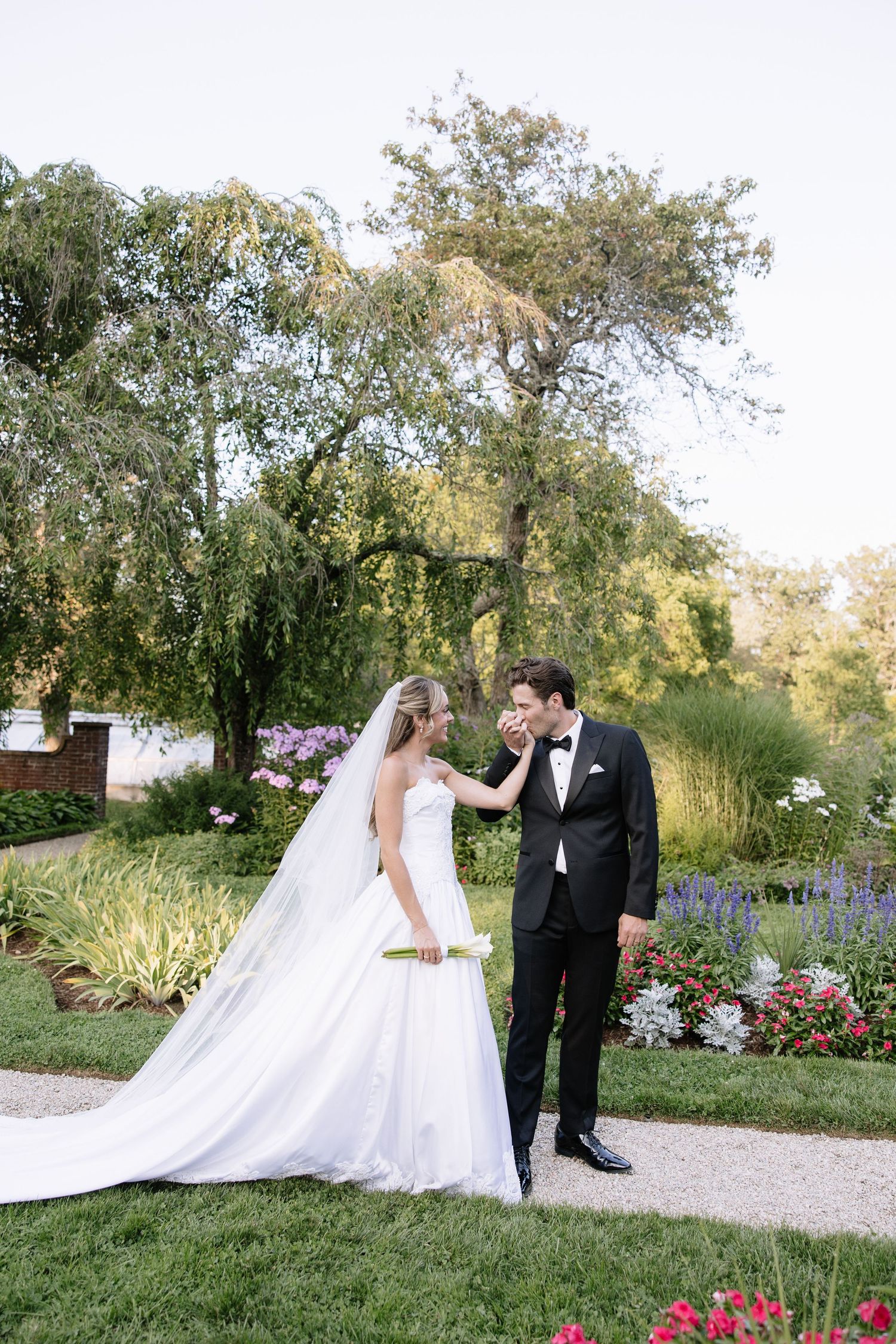 Wedding couple poses along garden path with flowing veil and floral landscaping.