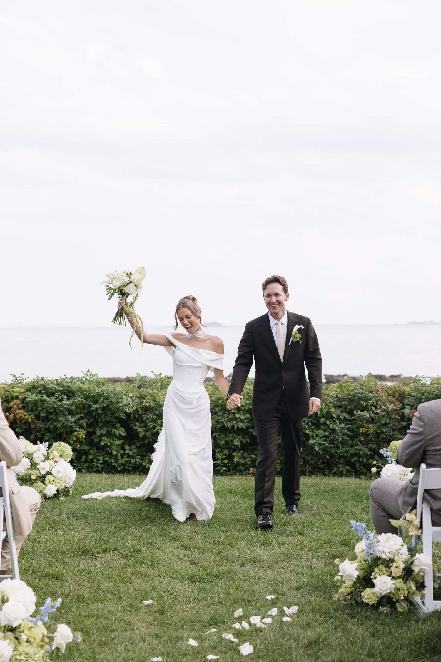 Joyful bride raising bouquet and groom walking down outdoor aisle after wedding ceremony by the ocean.