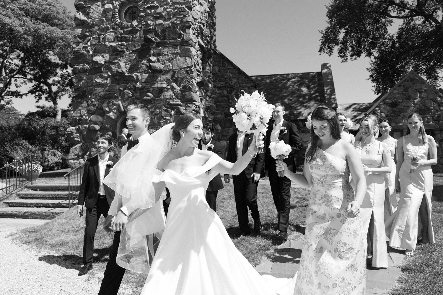 Black and white wedding photo of bride and groom walking with bridal party outside historic stone church.