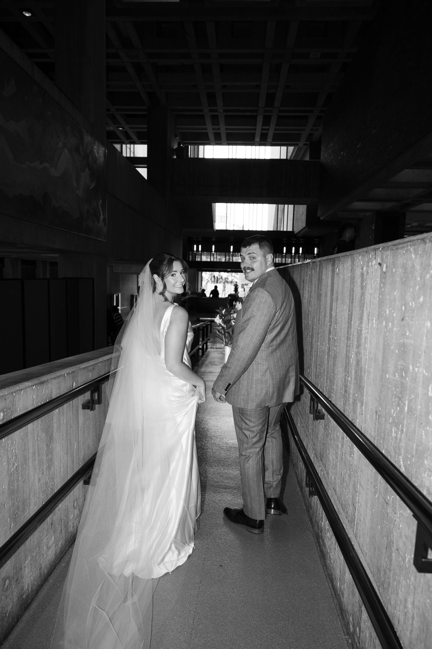 Black and white photo of a bride and groom holding hands on a walkway, smiling at each other on their wedding day.