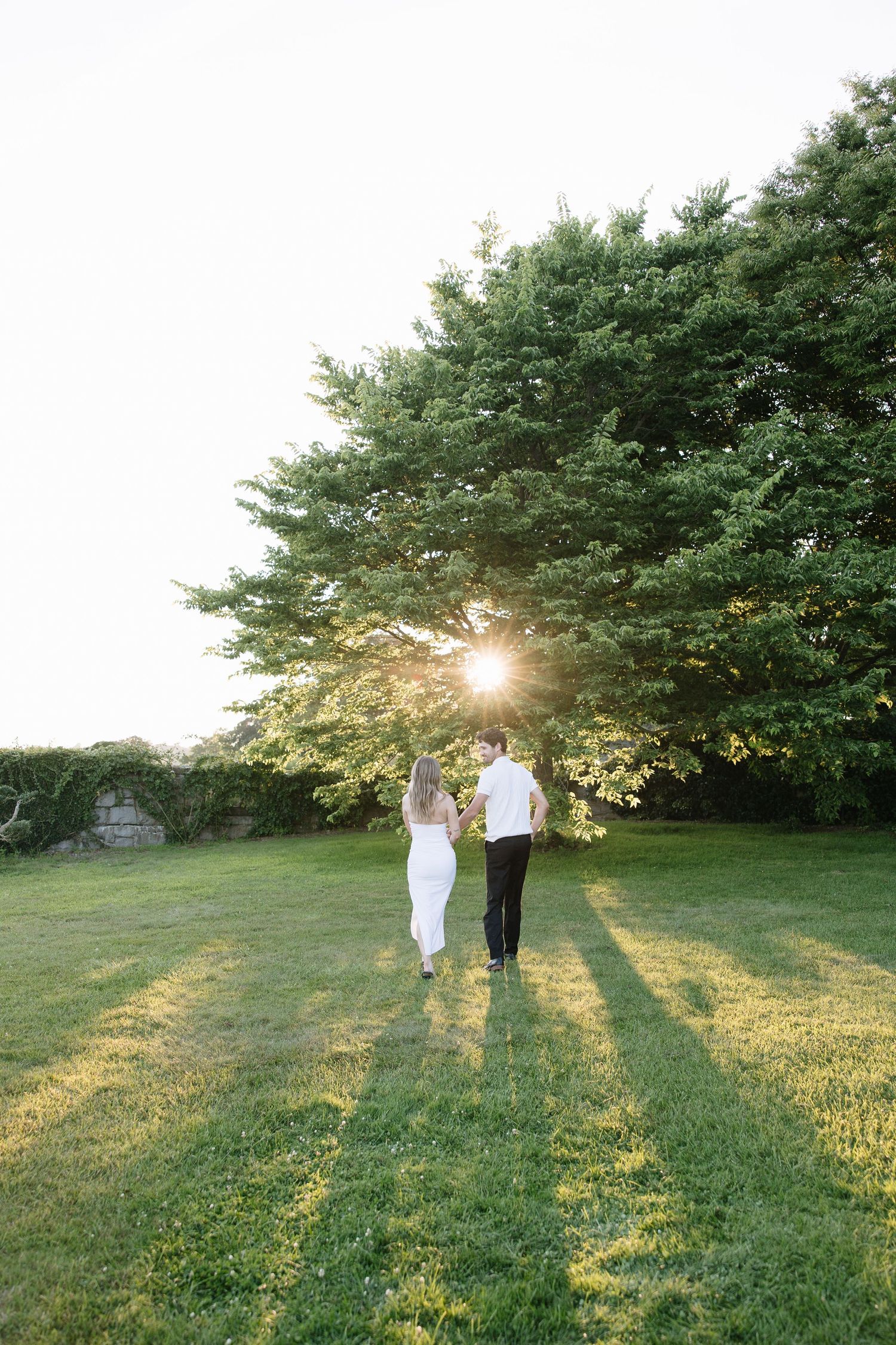 Couple holding hands walking toward sunset through lush green lawn with large trees glowing in golden light.