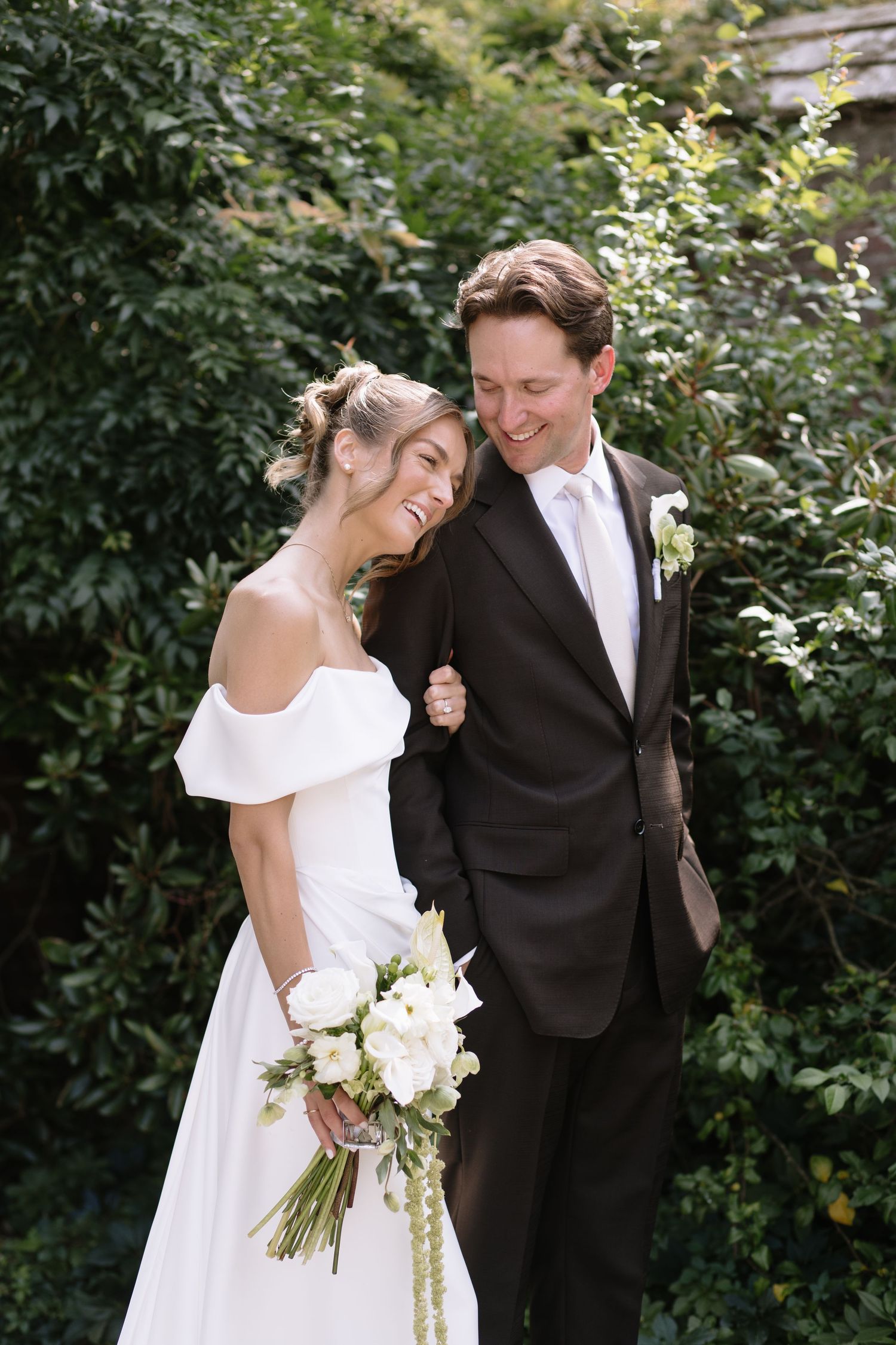 A smiling bride in an off-the-shoulder white gown holds a white bouquet while leaning on her groom in a brown suit in a garden.