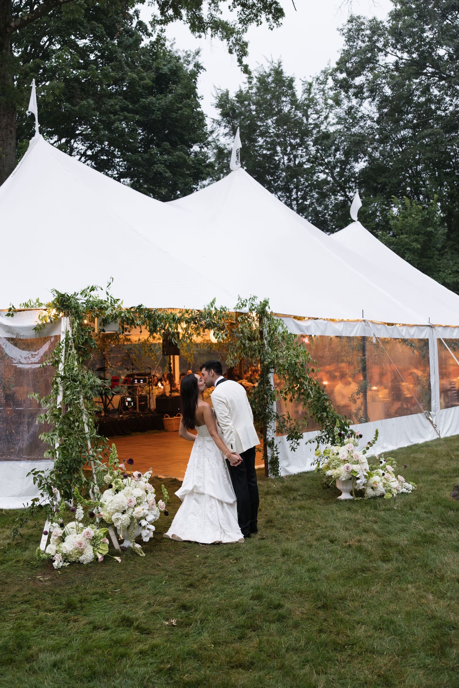 Bride and groom kiss beneath a floral arch at the entrance of a white sailcloth wedding tent at dusk.