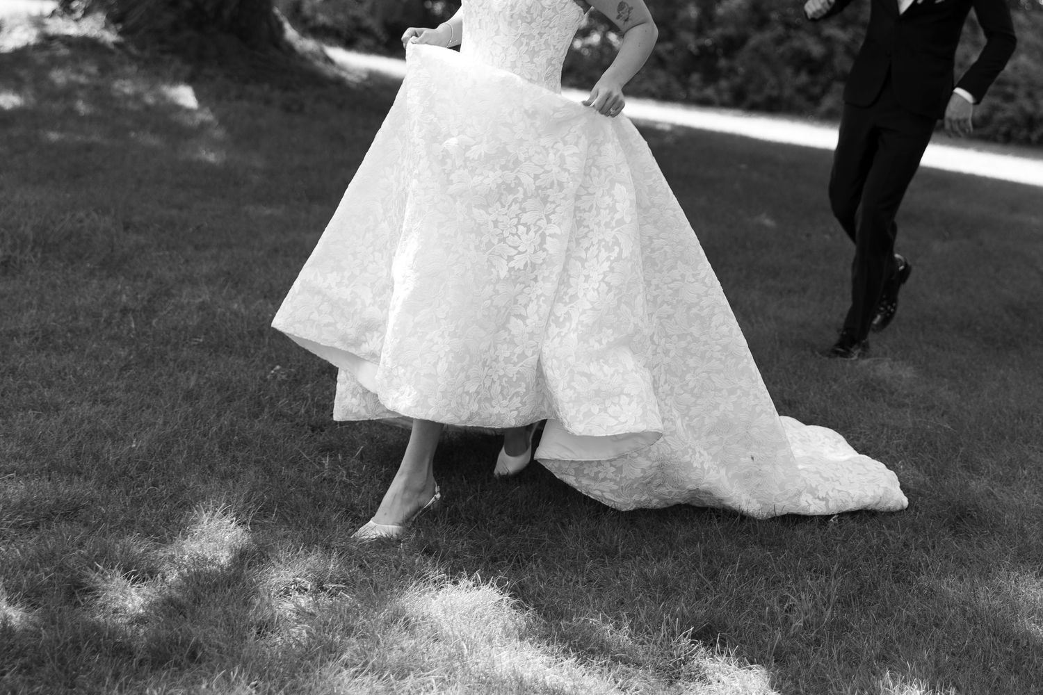 Black and white photo of a bride in a lace wedding gown walking on grass with a groom in a dark suit.