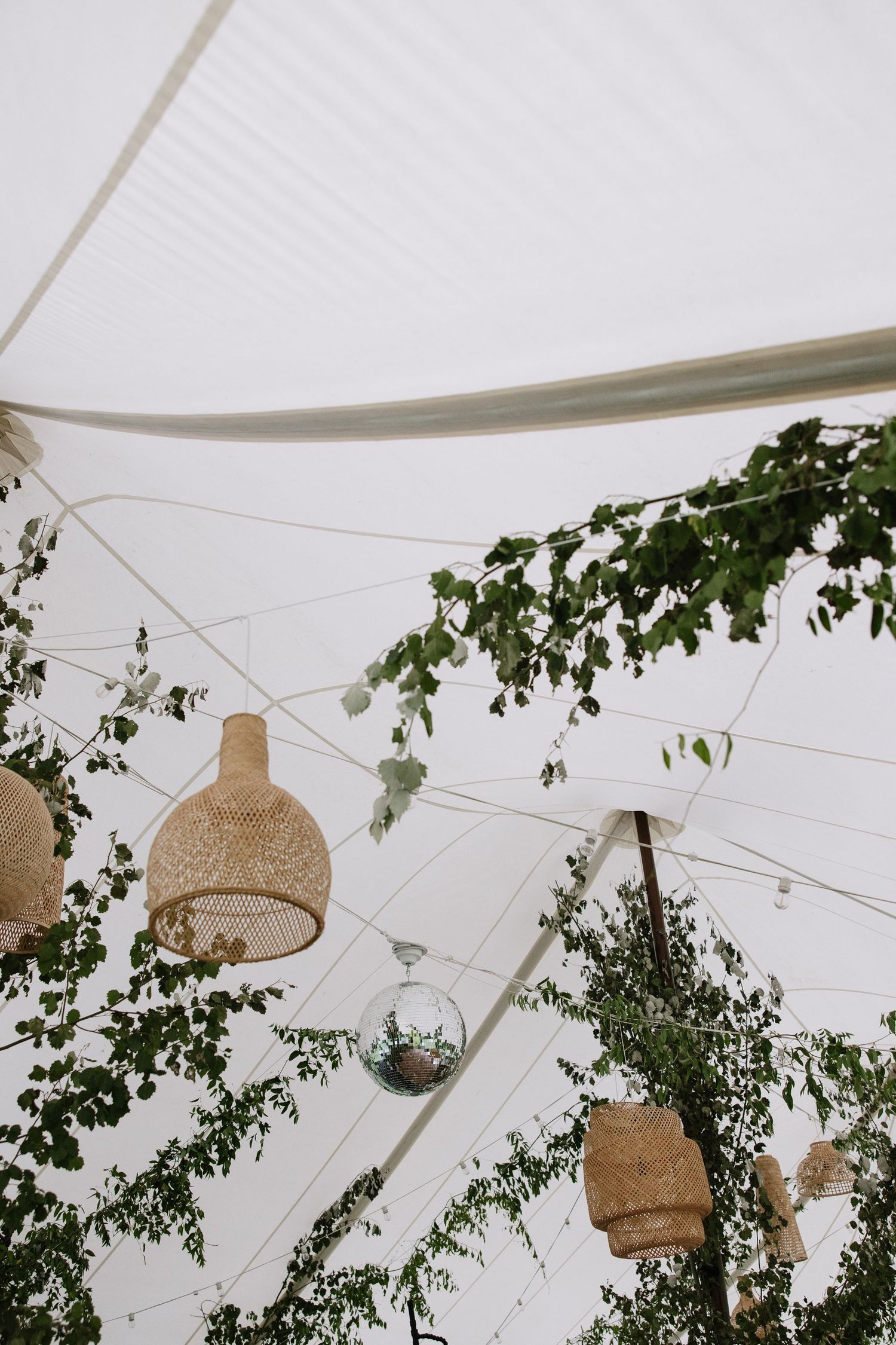Boho wedding tent ceiling decorated with wicker pendant lights, string lights, and lush green foliage branches.