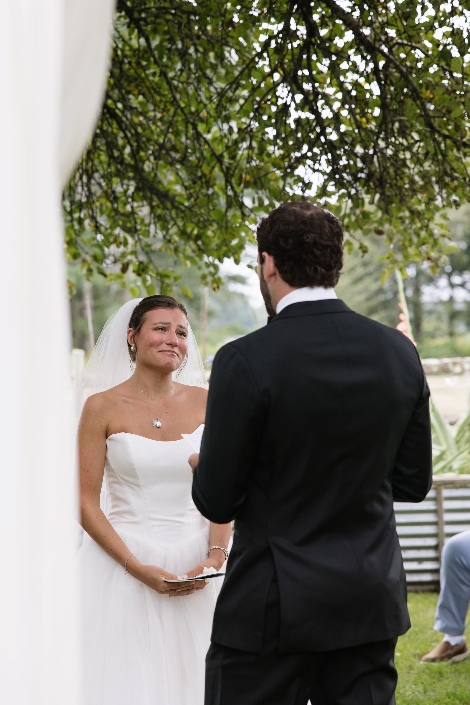 Emotional bride in white strapless gown crying during outdoor wedding ceremony while groom reads vows under green trees.