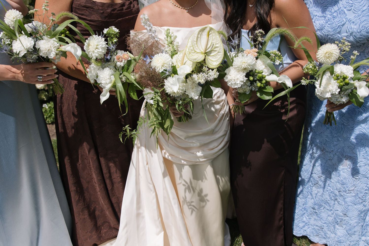 Bride in white gown holding white floral bouquet flanked by bridesmaids in brown dresses at outdoor wedding.