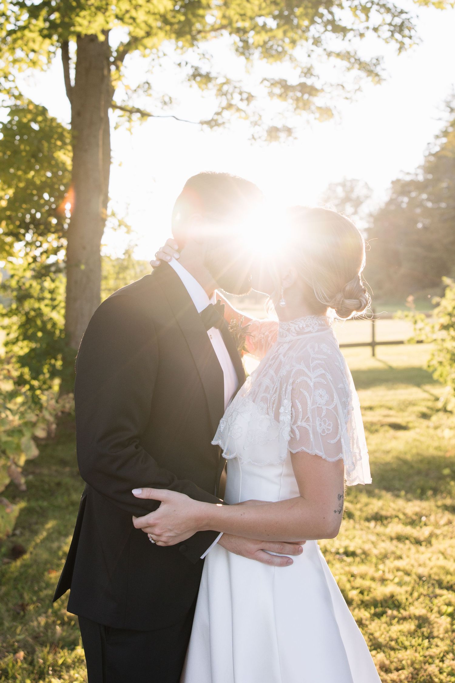 Bride and groom sharing a kiss outdoors at golden hour with sunlight glowing behind them in a lush green setting.