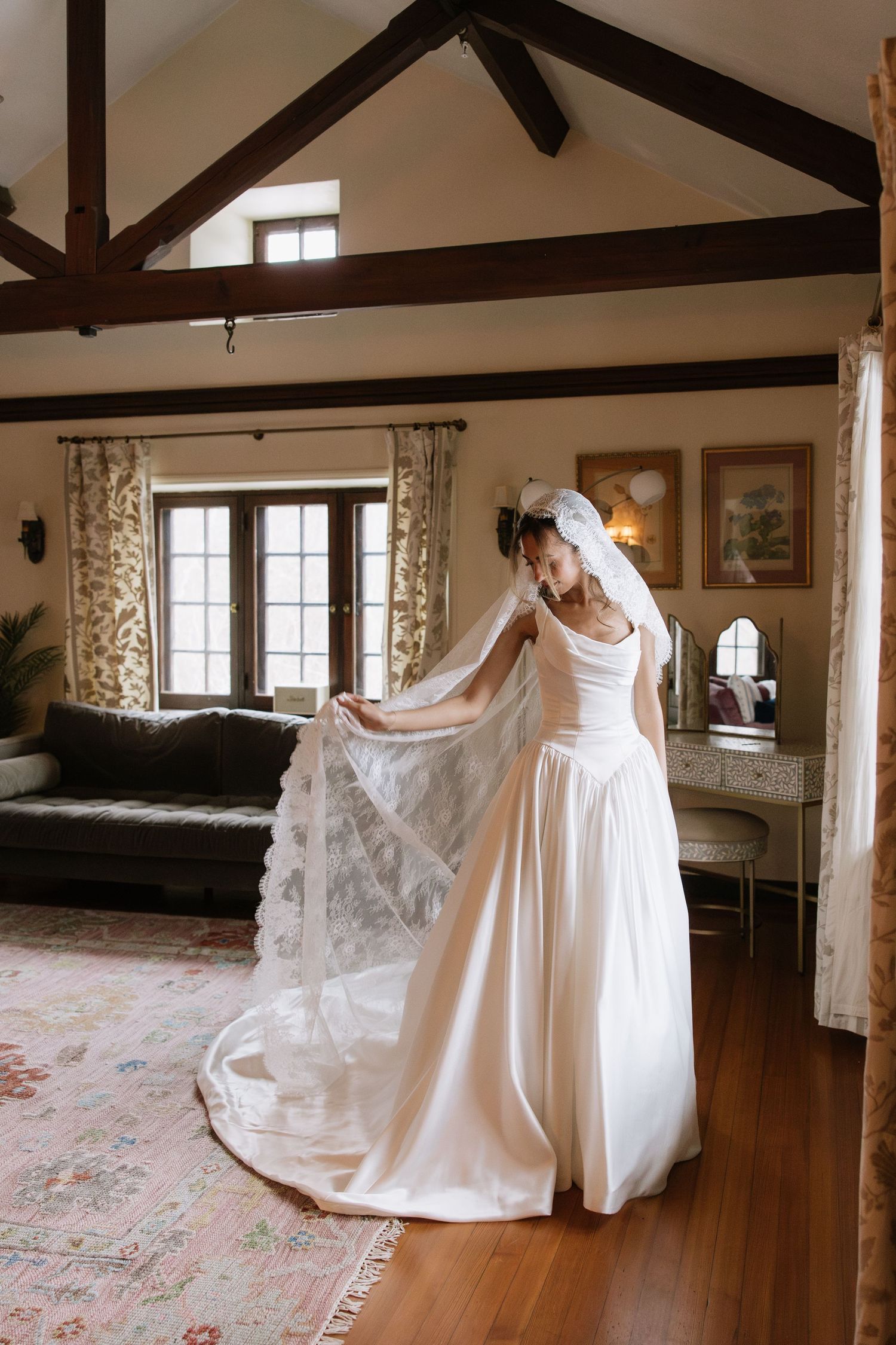 A flowing white wedding dress with delicate lace veil in a rustic room with exposed wooden beams and floral curtains.