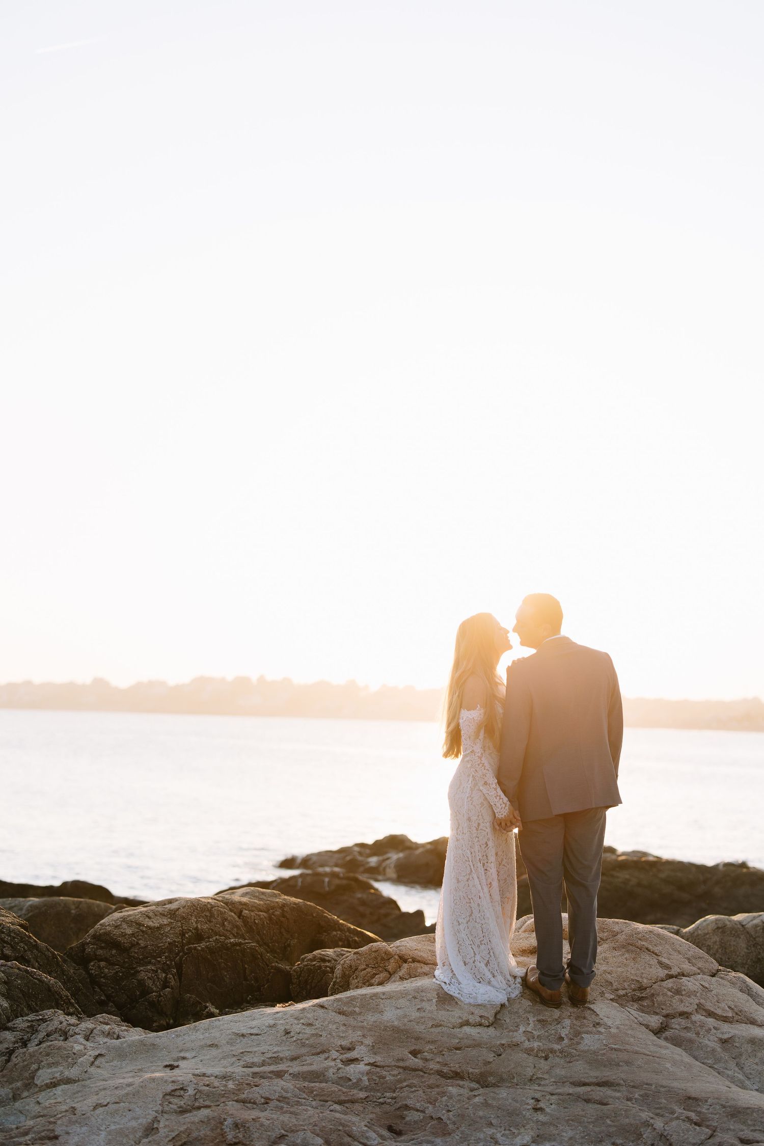 Couple standing together on rocky shore overlooking calm waters at sunset.