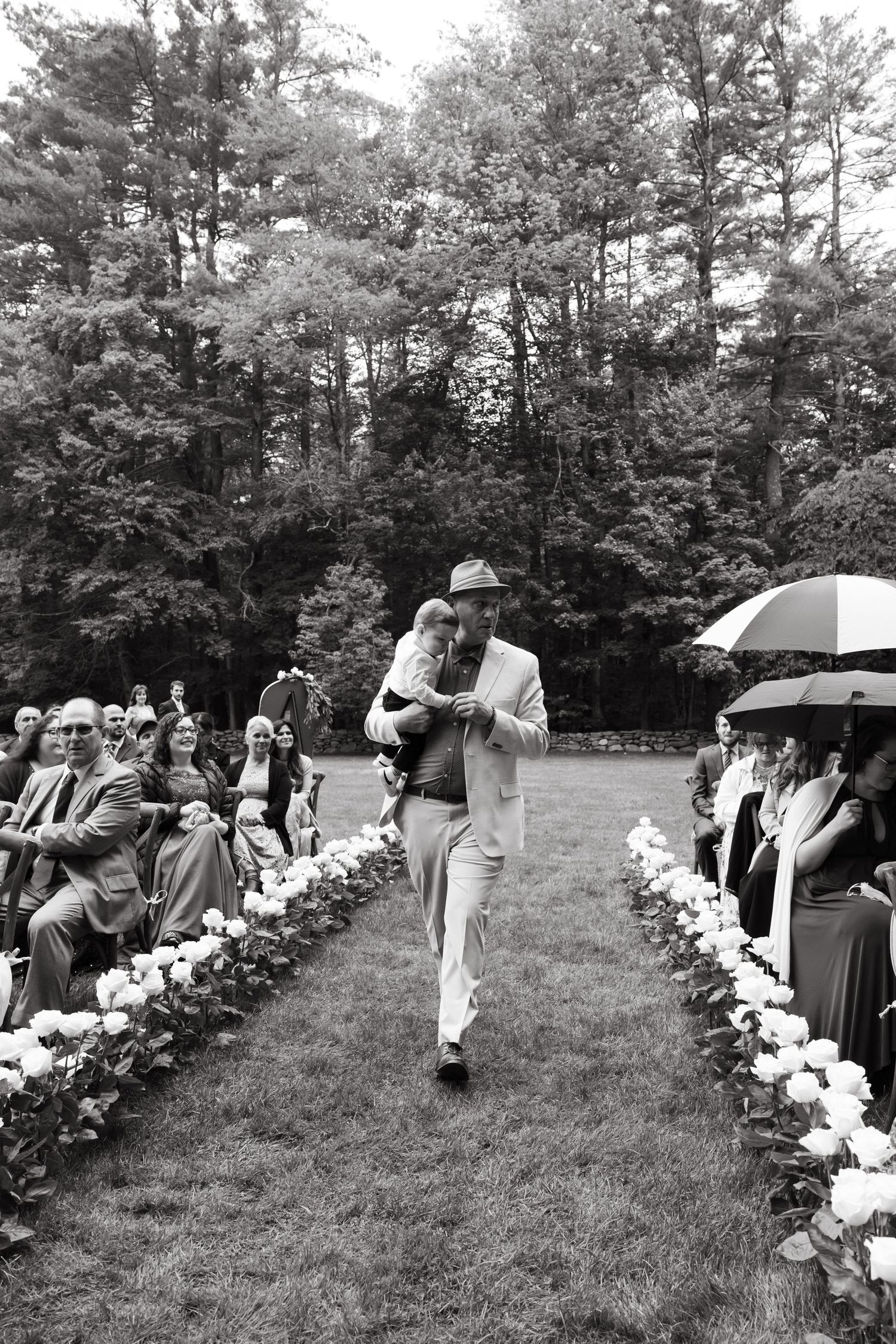 Black and white photo of wedding guests with umbrellas lining a flower-decorated aisle.