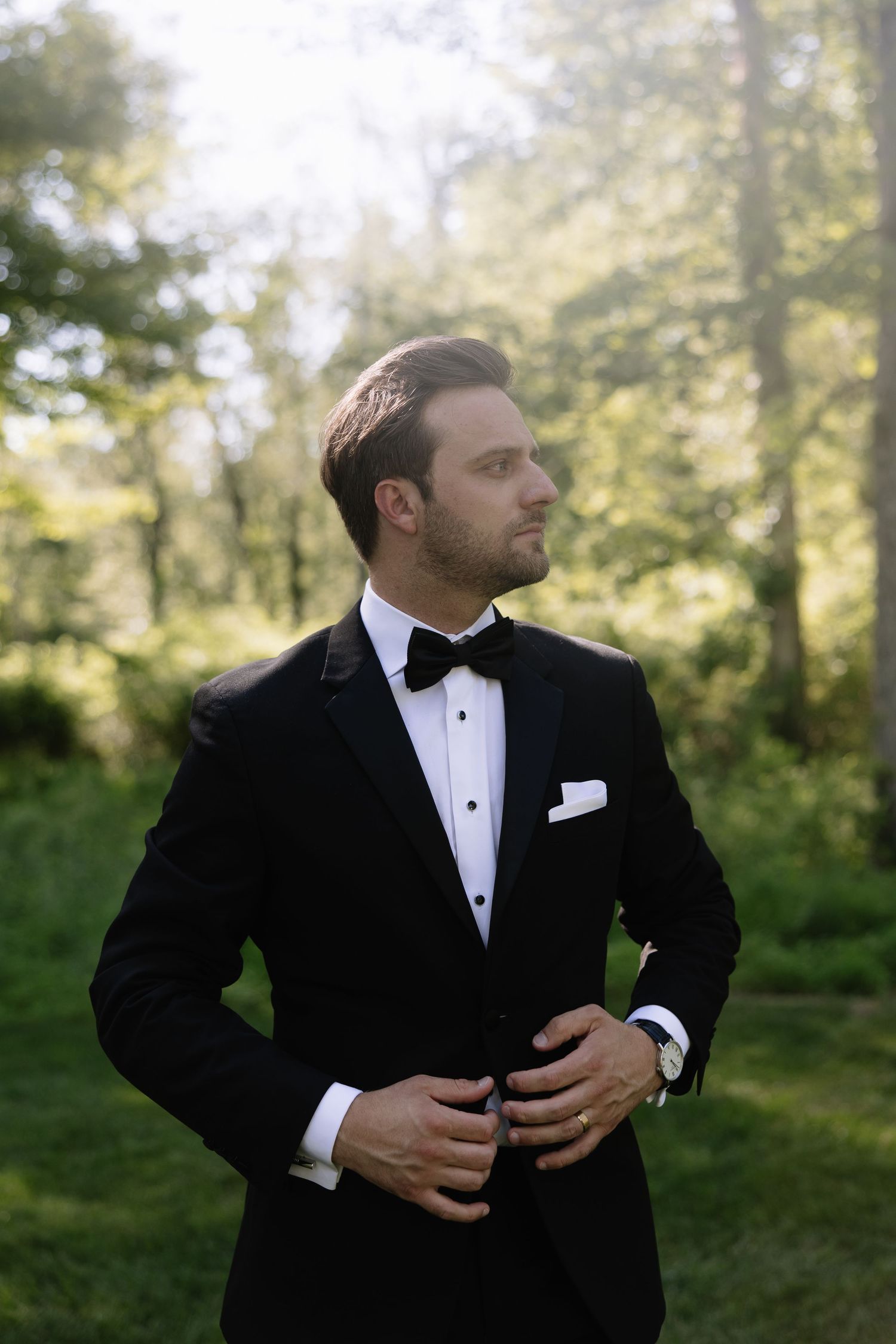 Groom in classic black tuxedo adjusts his jacket in outdoor wedding setting.