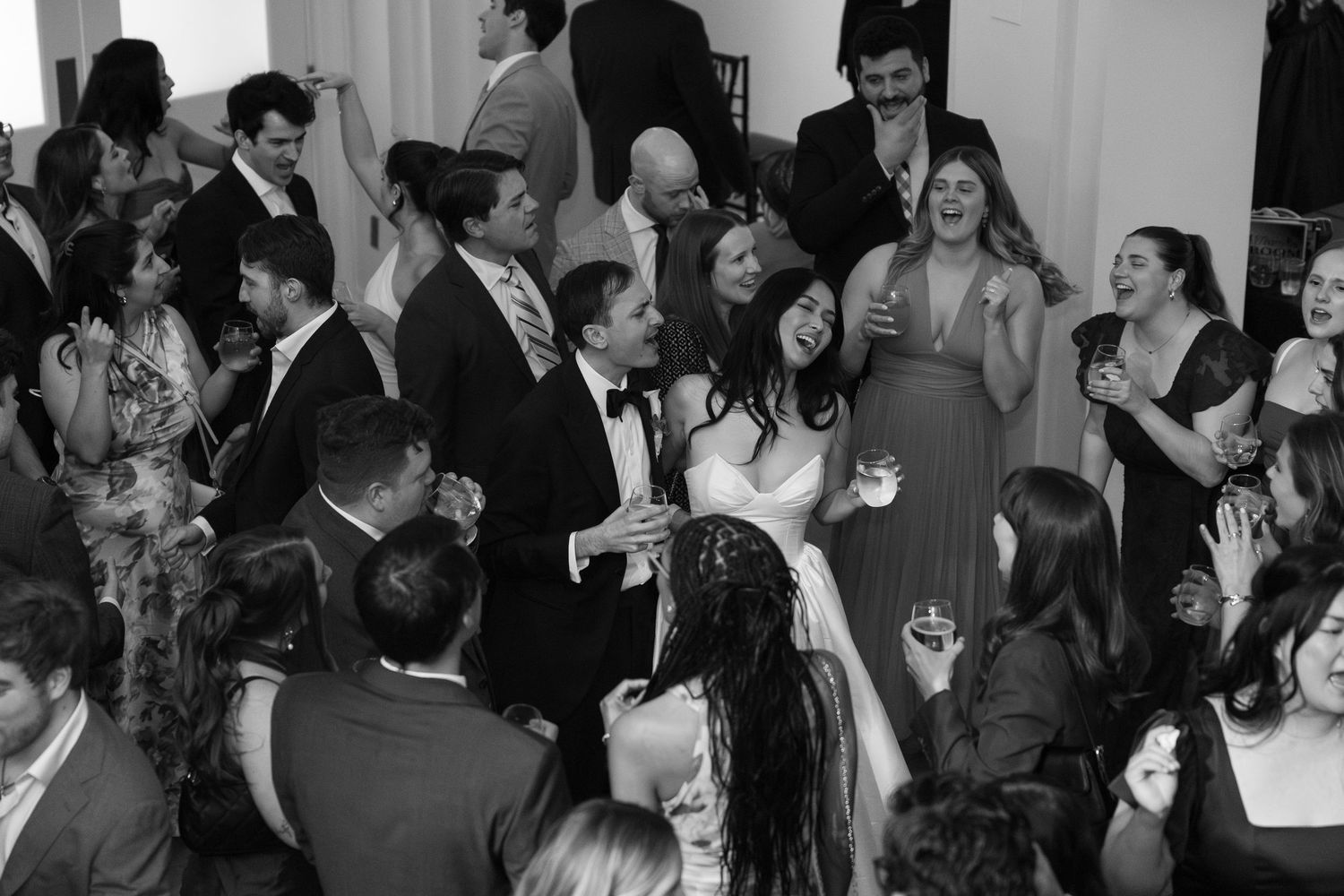 Black and white photo of bride and groom laughing with wedding guests on a crowded dance floor.