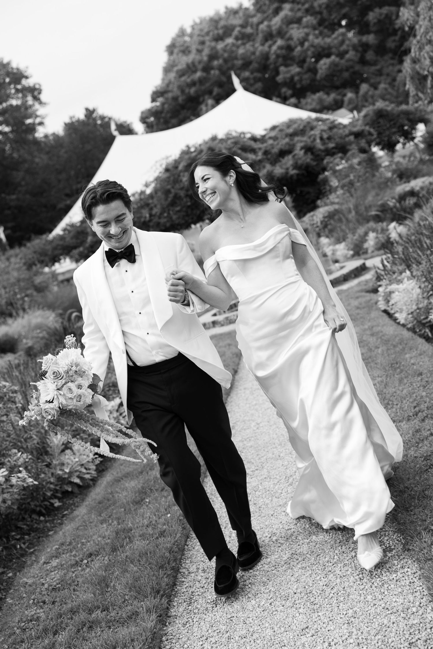 Black and white photo of smiling bride and groom walking arm in arm on garden path, bride in off-shoulder gown, groom in white tuxedo.
