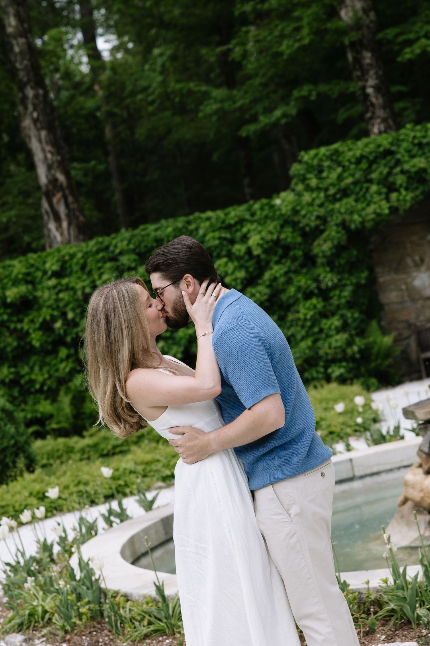 Couple sharing romantic kiss near stone fountain with white tulips and lush green backdrop.