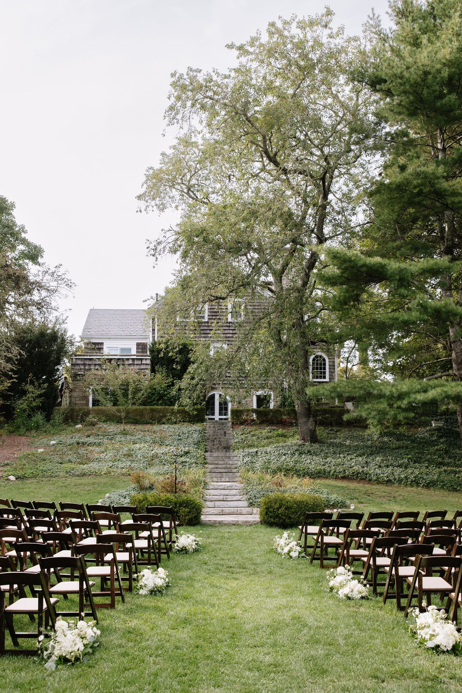 Outdoor wedding ceremony setup with dark wooden chairs on green lawn facing a charming stone estate house.