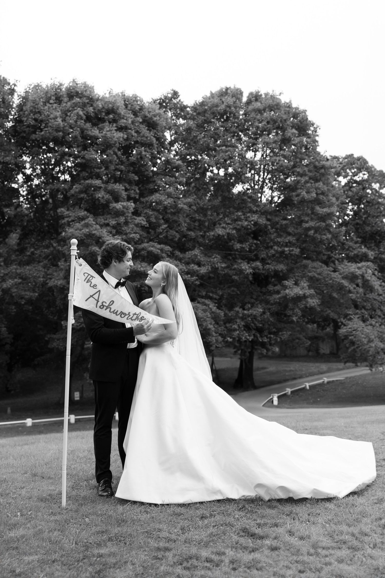 Black and white photo of bride and groom holding a personalized flag outdoors on their wedding day.