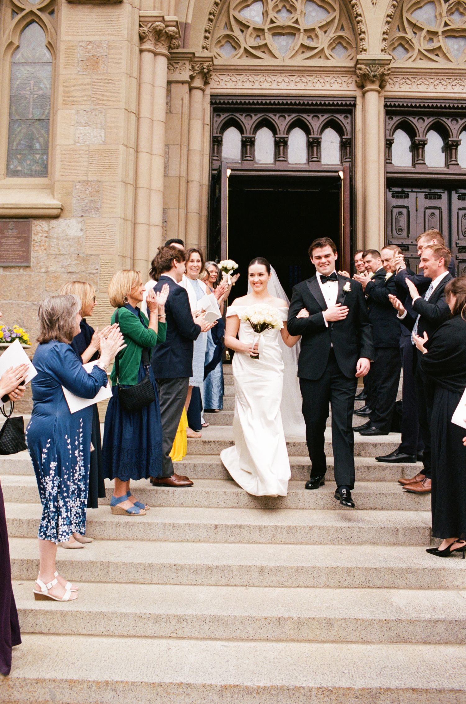 Newlywed couple exits a grand stone church, greeted by applauding guests lining the steps after their wedding ceremony.