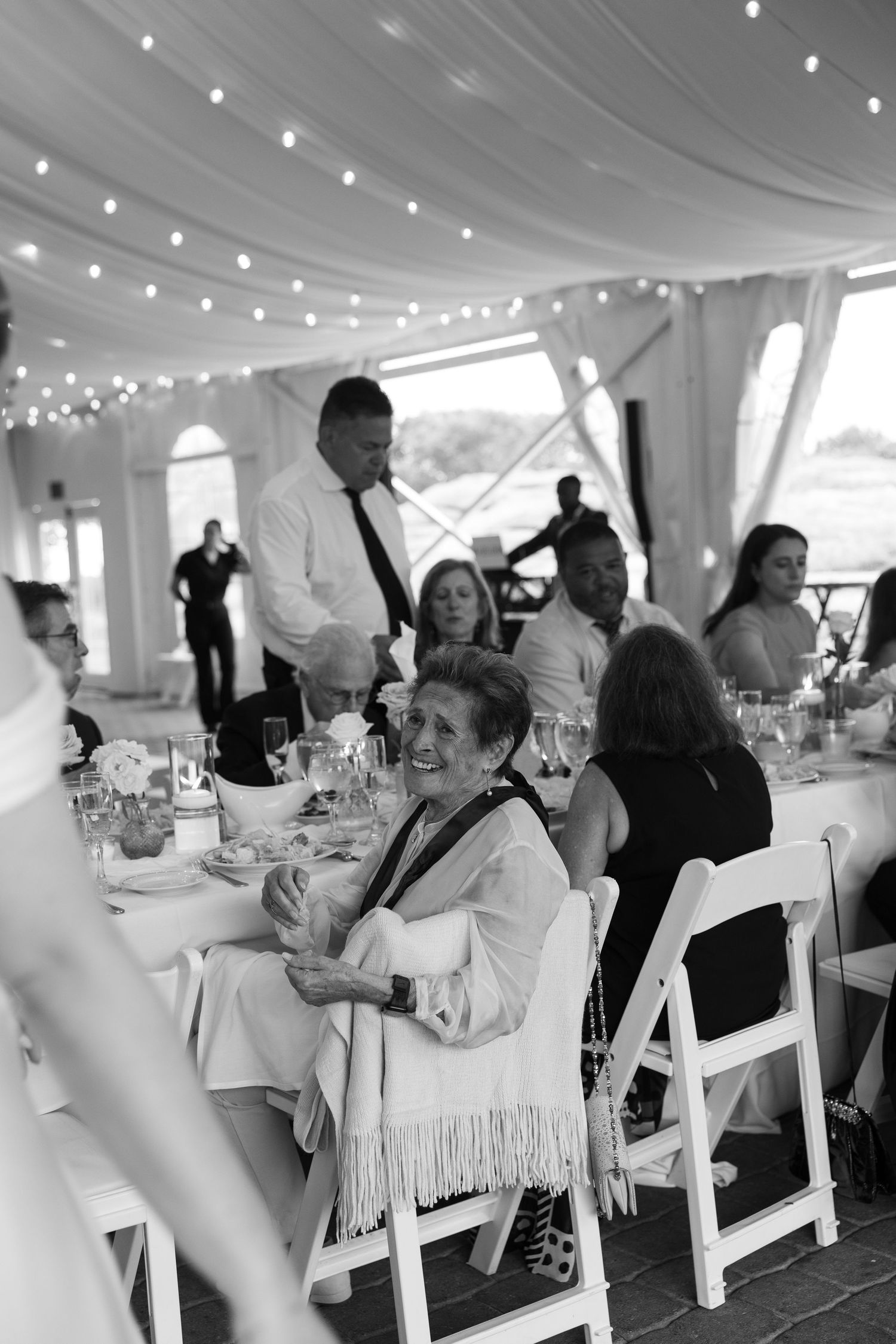 Guests laughing and celebrating at elegant wedding reception under white tent with string lights in black and white.