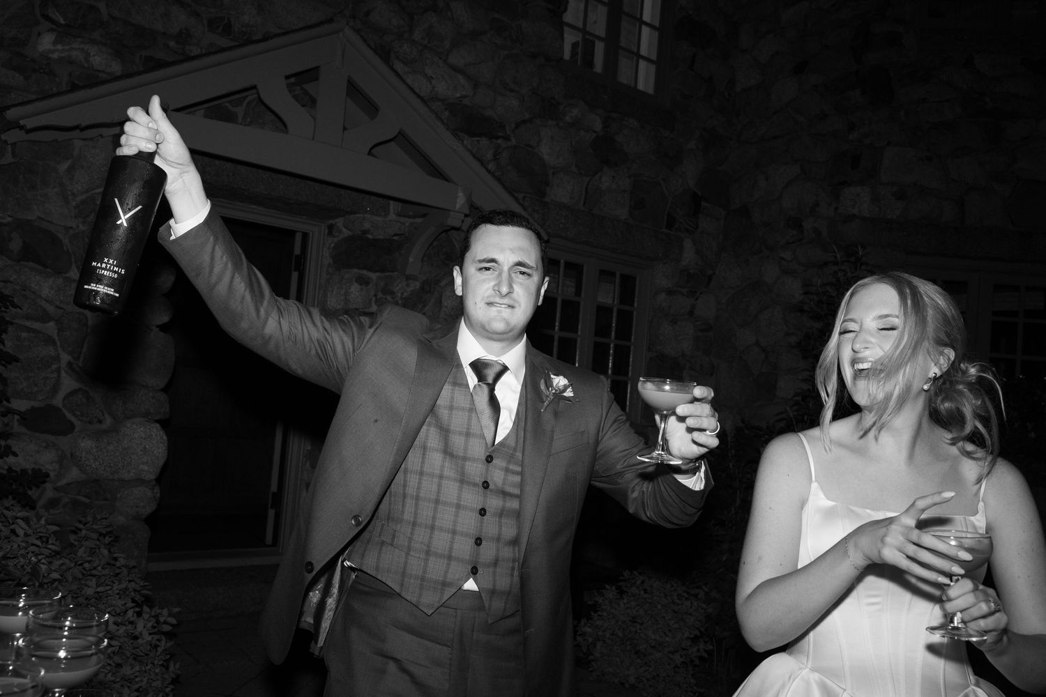 Groom raises champagne bottle while bride holds cocktail glass outdoors at night wedding celebration.
