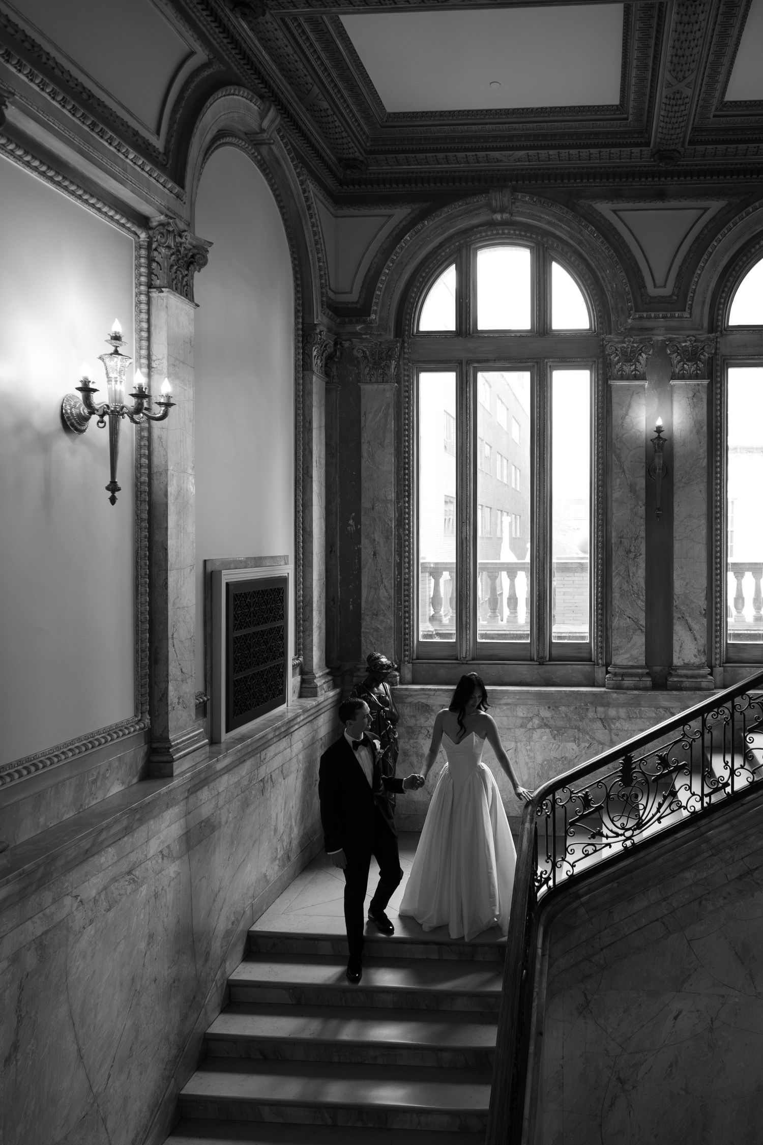Black and white photo of bride and groom walking hand in hand up grand marble staircase in ornate venue.