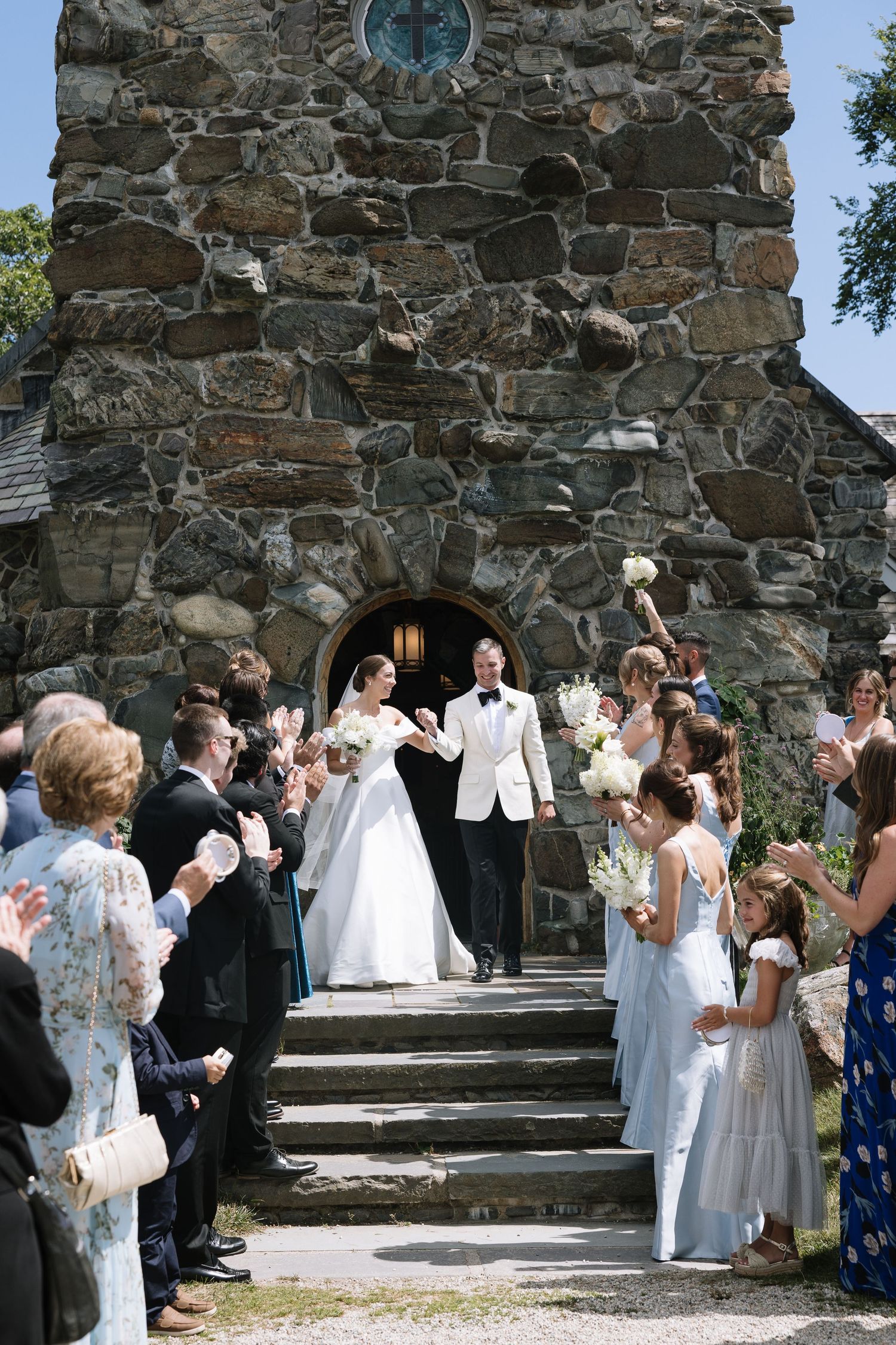 Bride and groom exit stone church surrounded by cheering guests on sunny wedding day.