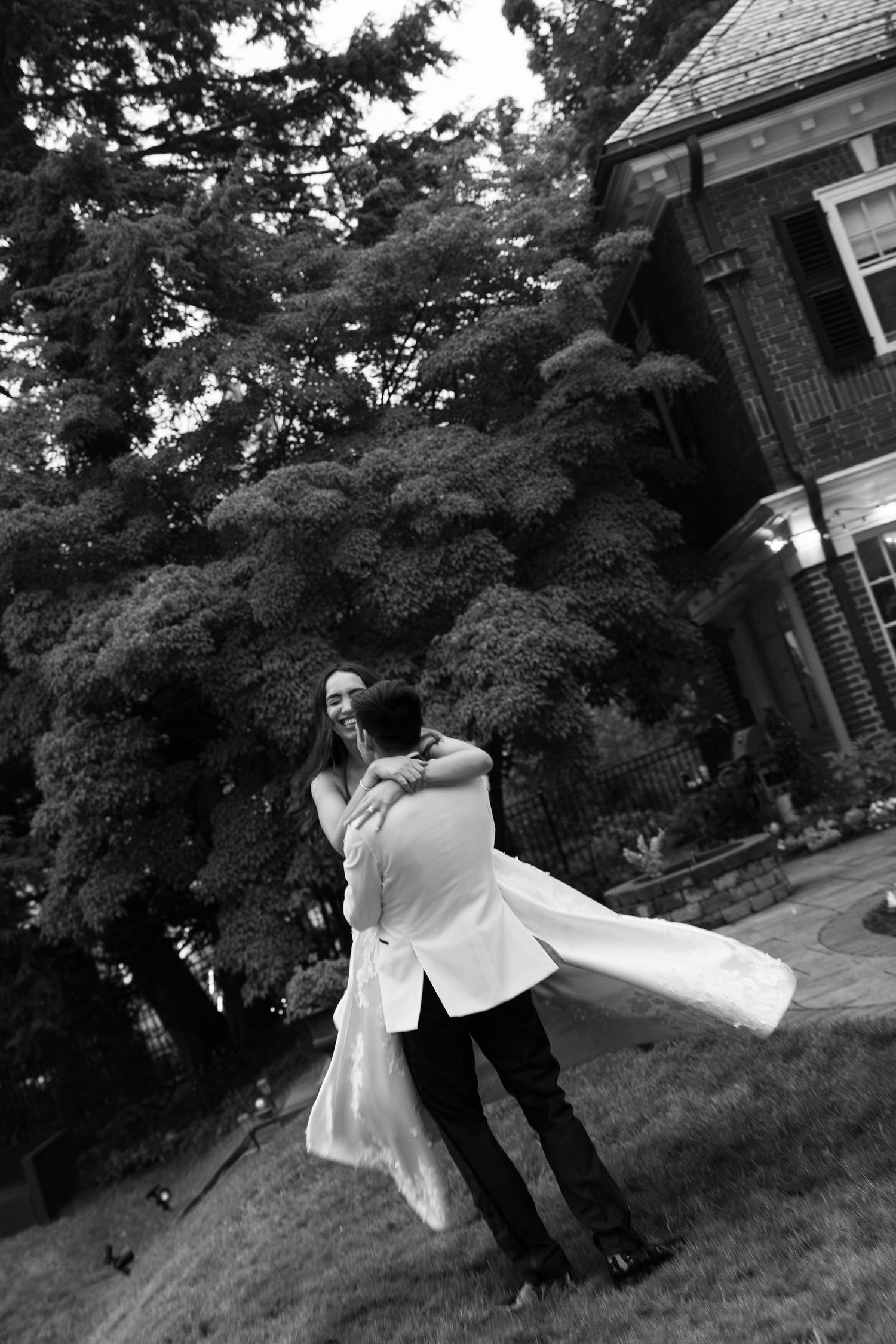 Black and white photo of groom lifting bride in wedding dress outdoors near brick estate with large trees.