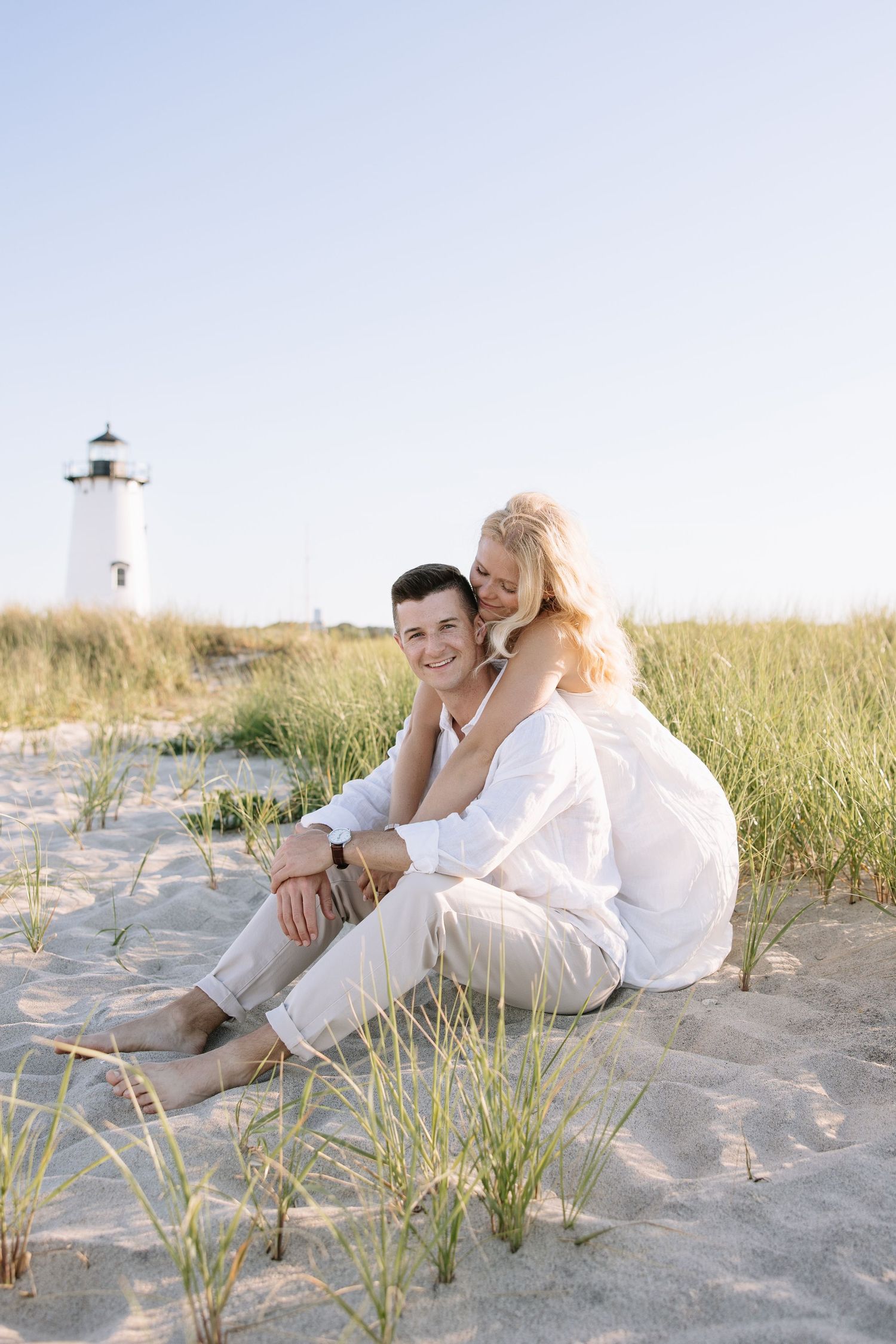 Couple sitting on sandy beach dunes in white outfits, embracing near a lighthouse on a sunny day.