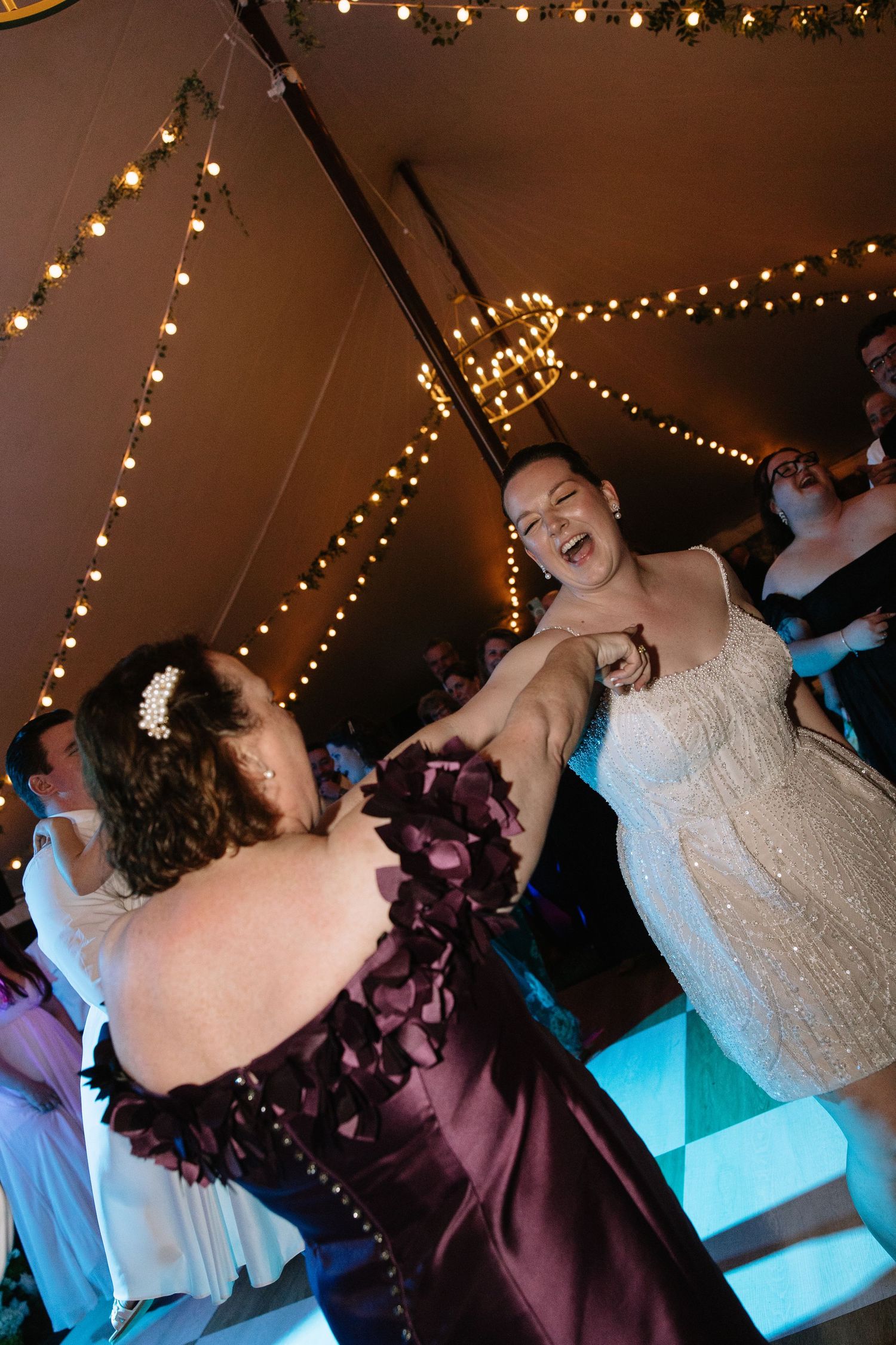 Two women dancing joyfully at a wedding reception under warm string lights in a decorated tent venue.