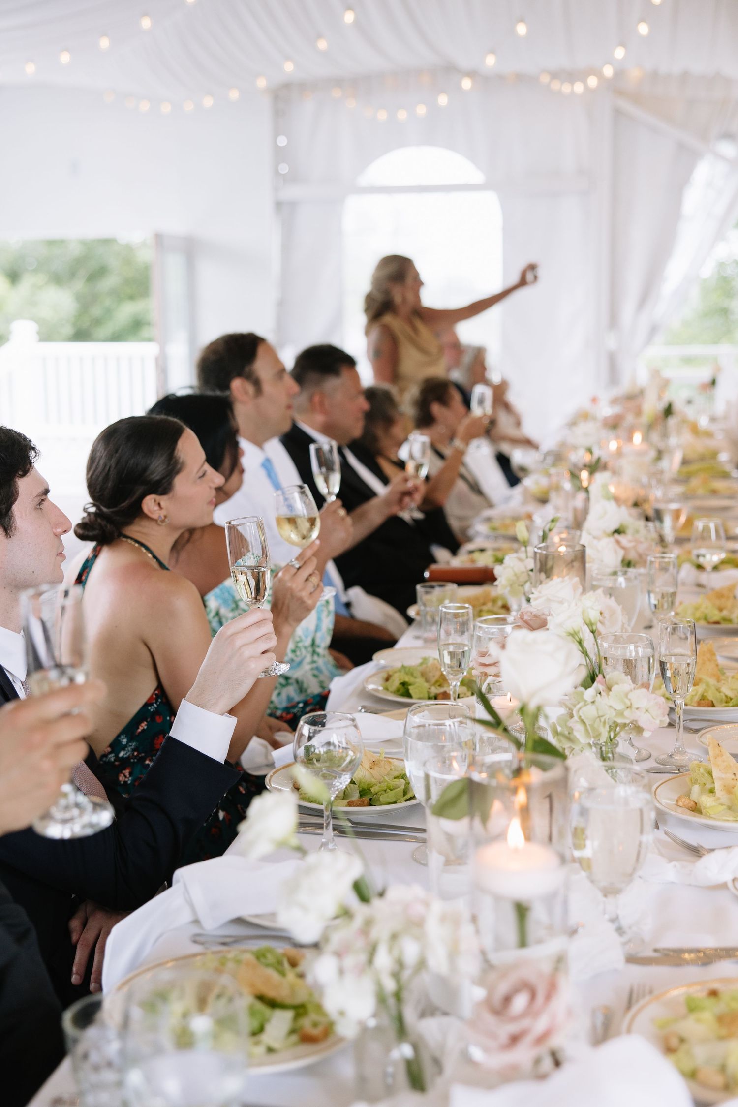 Wedding reception guests raise glasses in a toast at an elegantly decorated table under white draping.