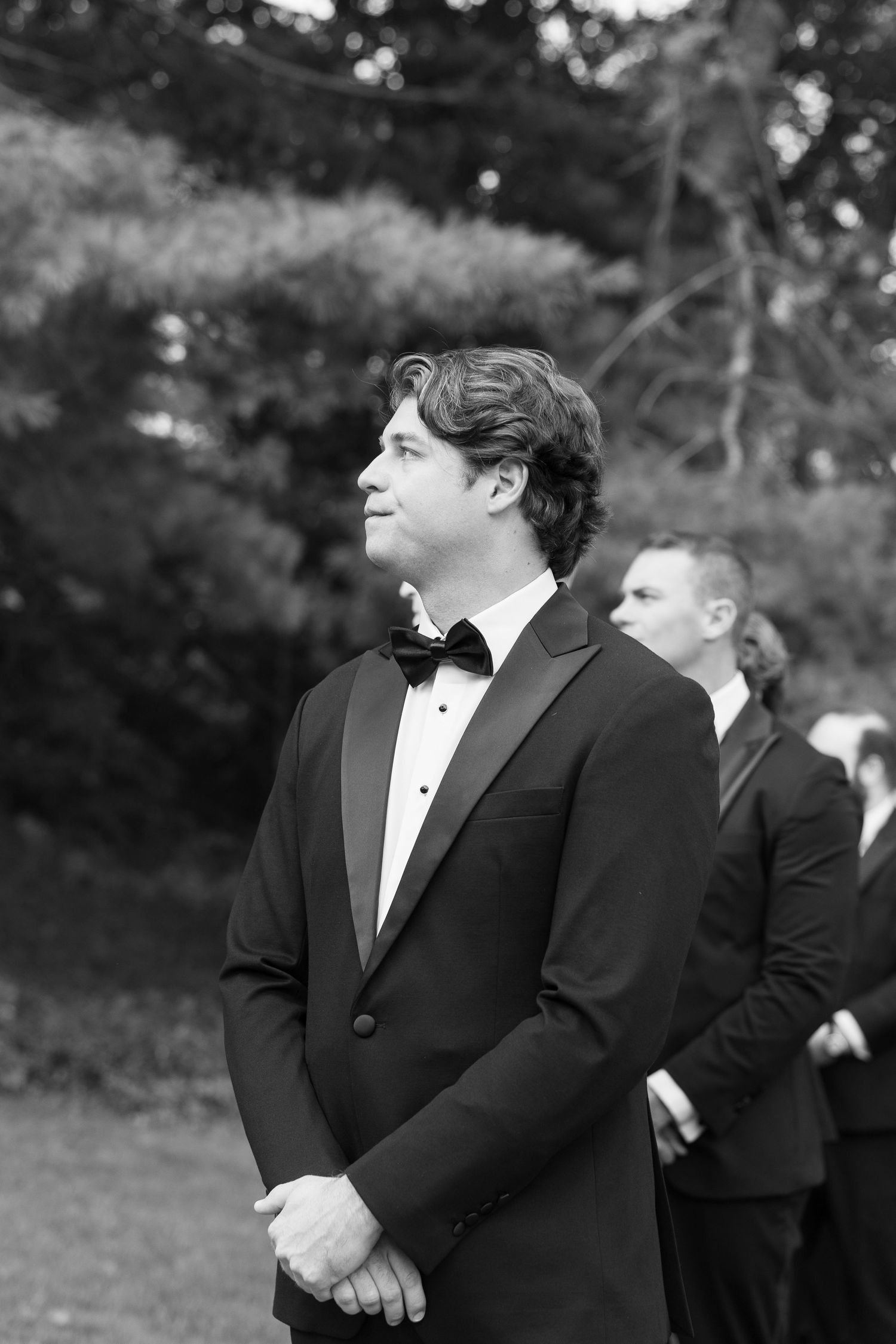 Groomsman in classic black tuxedo and bowtie stands at attention during outdoor ceremony.