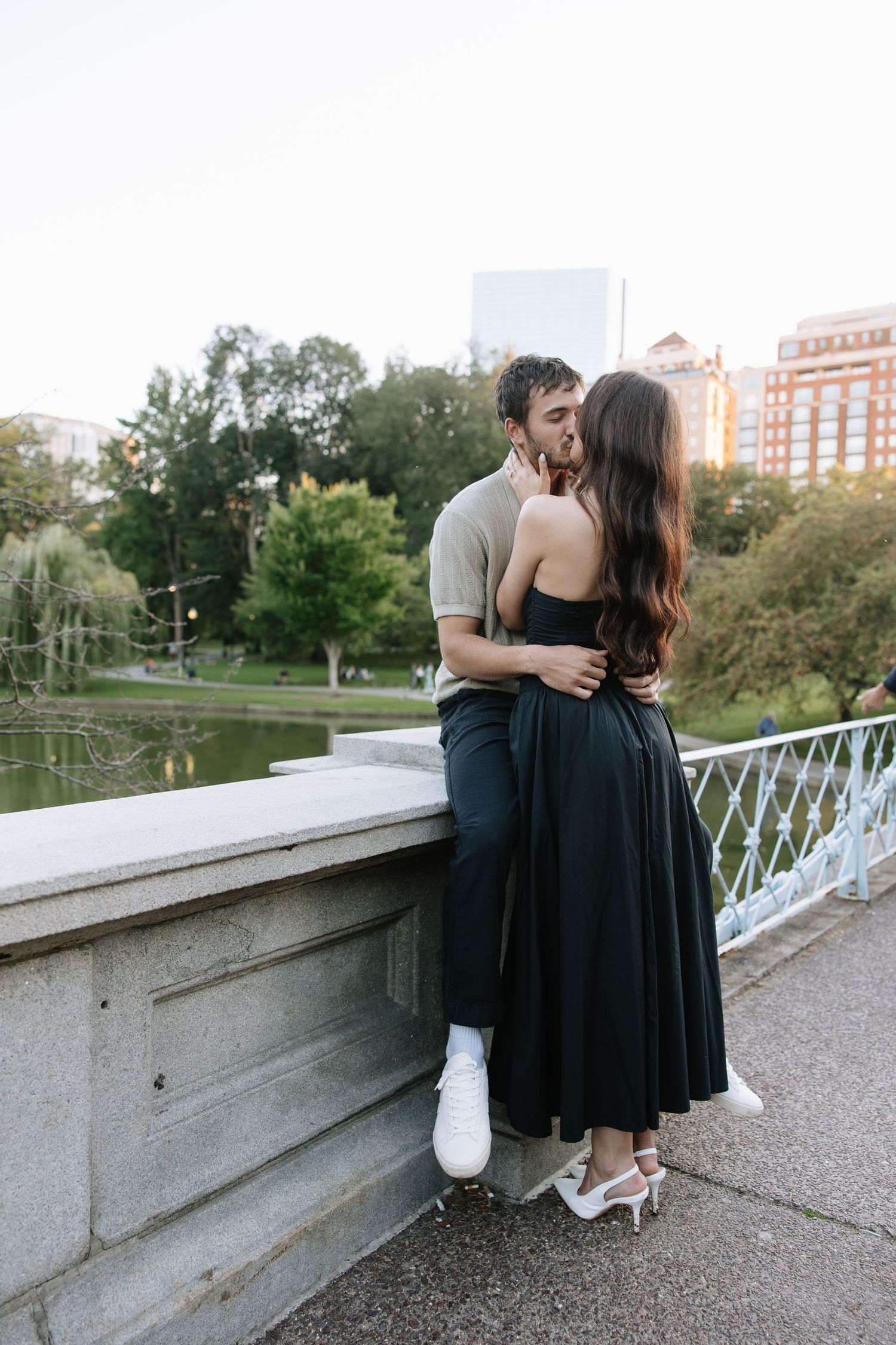 A couple leans against a concrete bridge railing in an urban park setting.