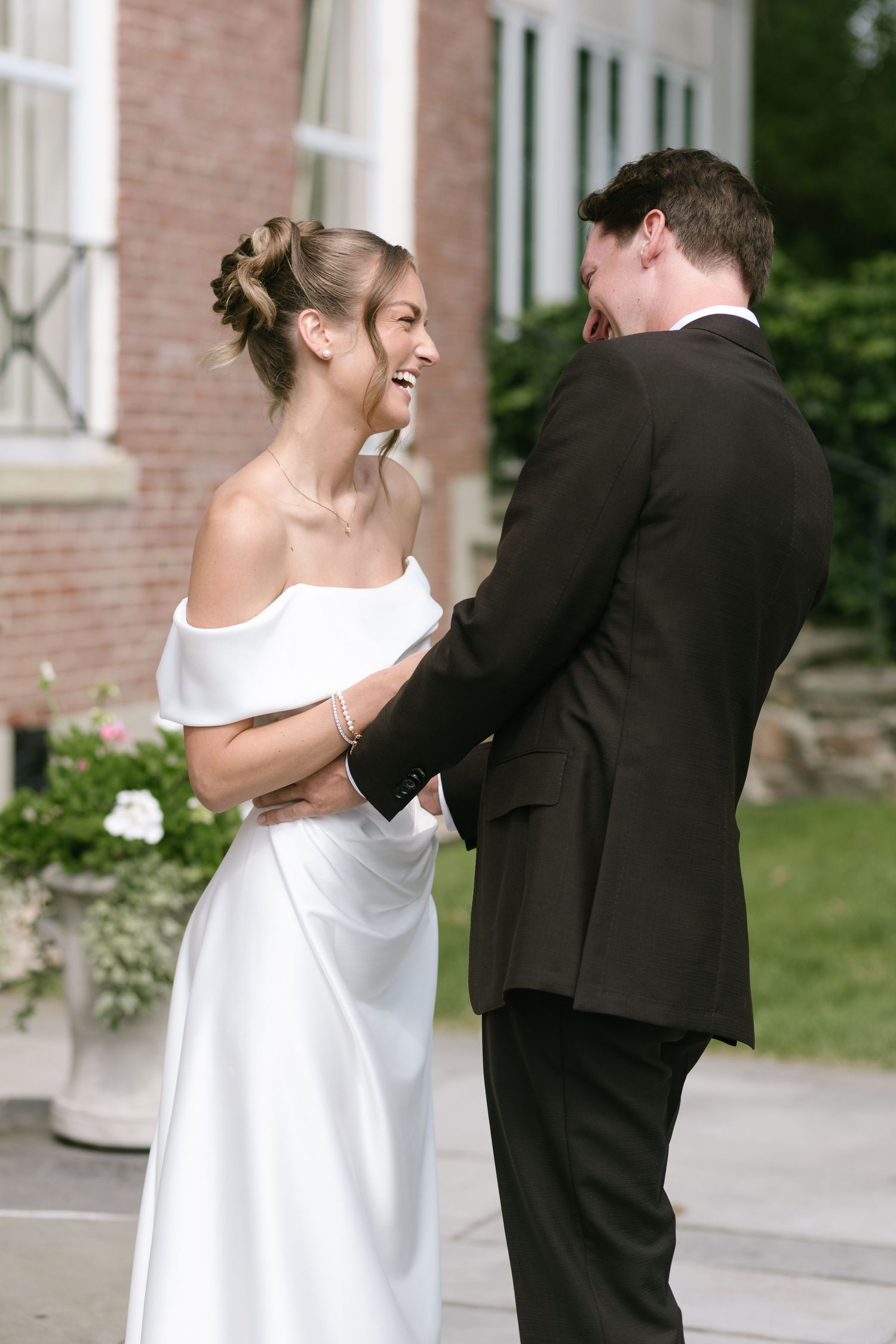 A smiling bride in an off-the-shoulder white satin gown embraces her groom in a dark suit outside a brick building on their wedding day.
