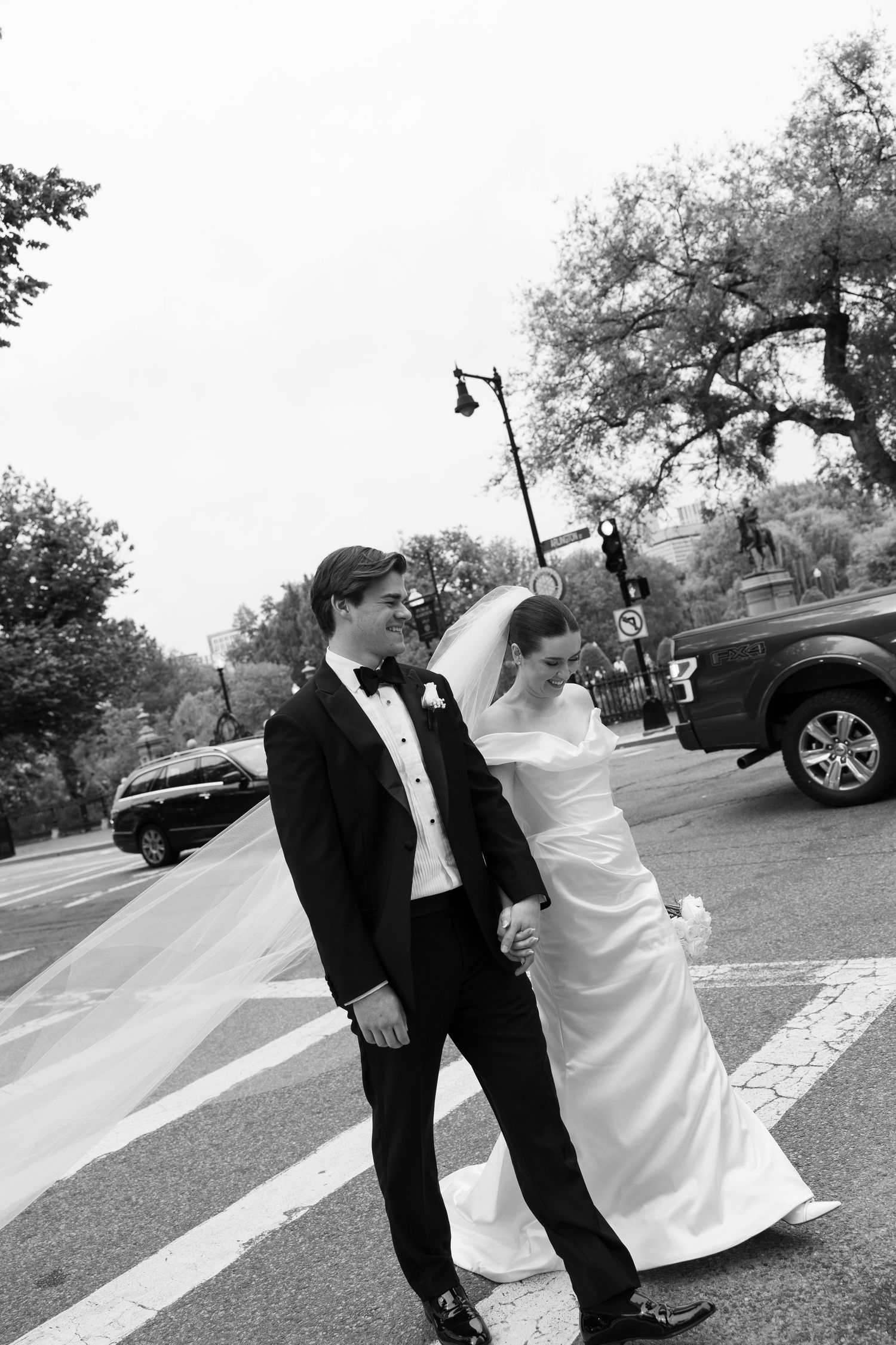 Bride and groom cross a city street in black and white, bride wearing off-shoulder gown with flowing veil.