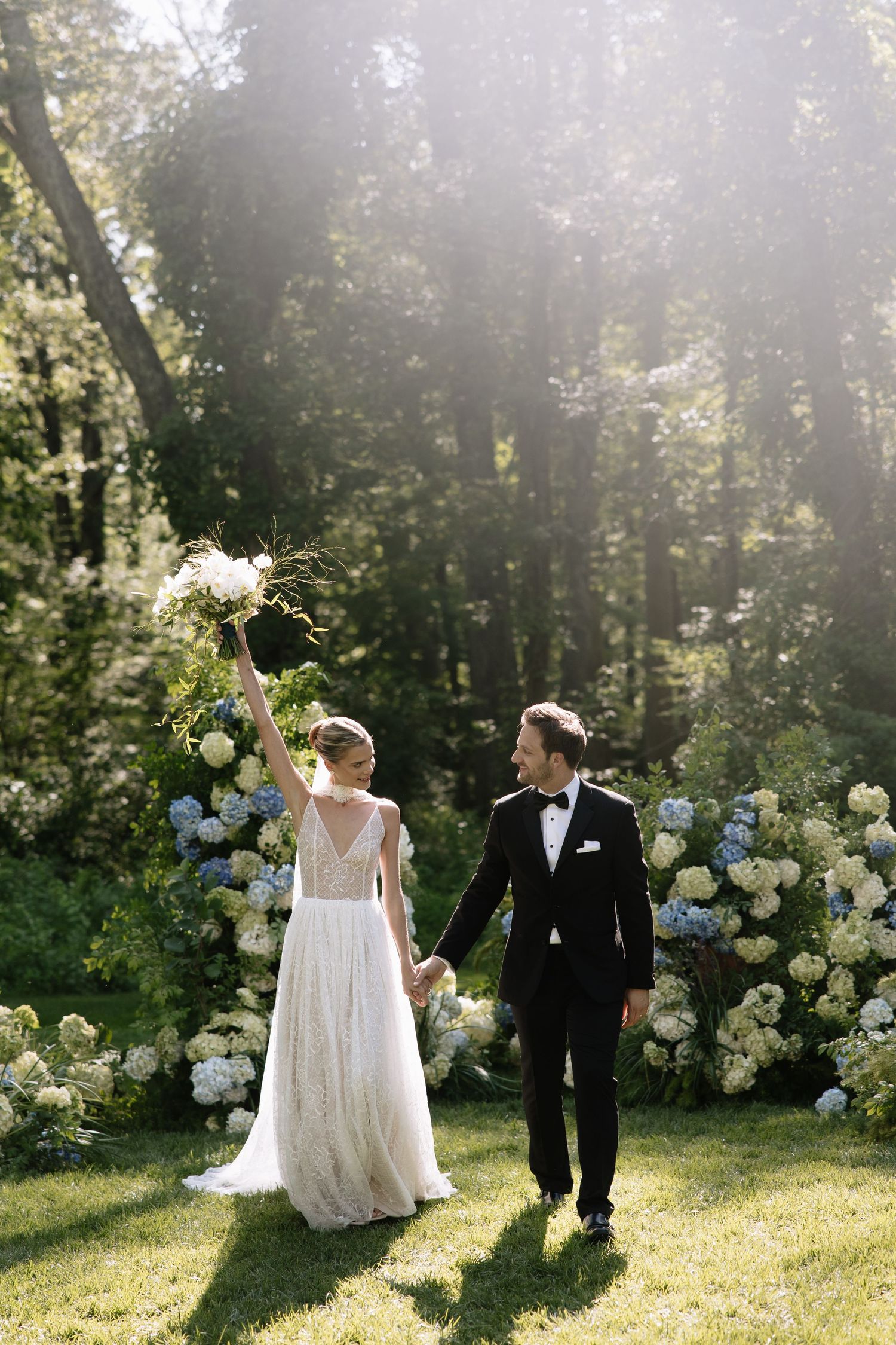 Bride raises bouquet while groom holds her hand at outdoor wedding ceremony with white and blue hydrangeas.