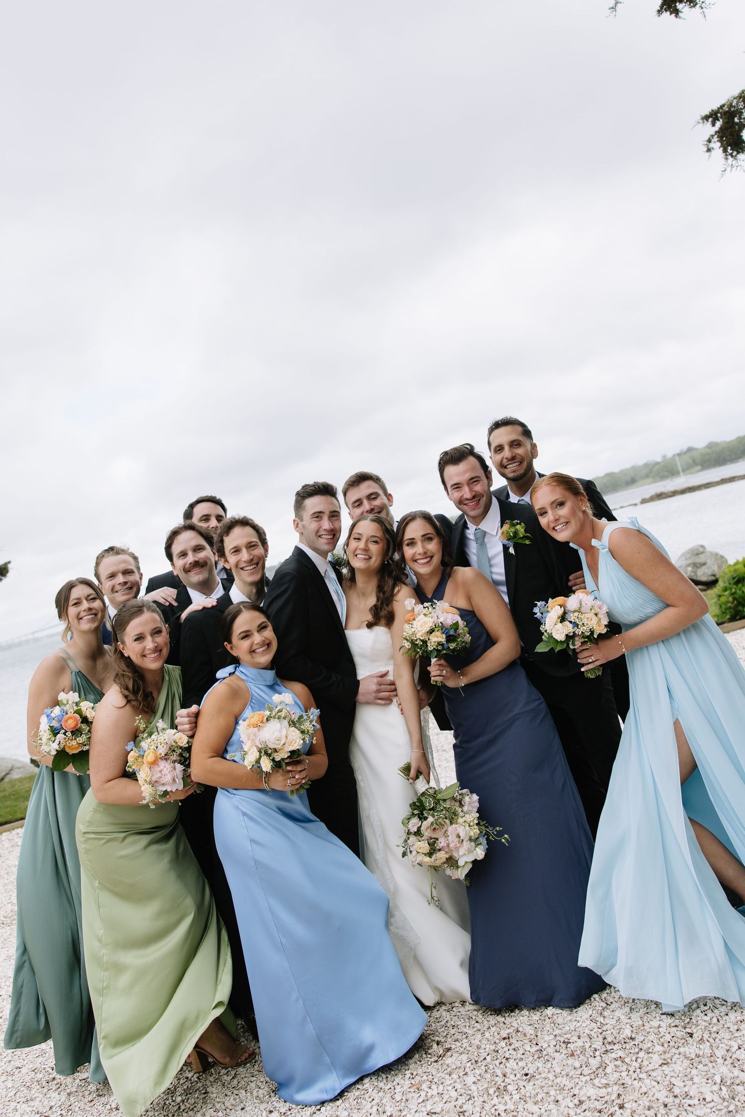 Wedding party in blue and green gowns poses with bride and groom outdoors near waterfront venue.