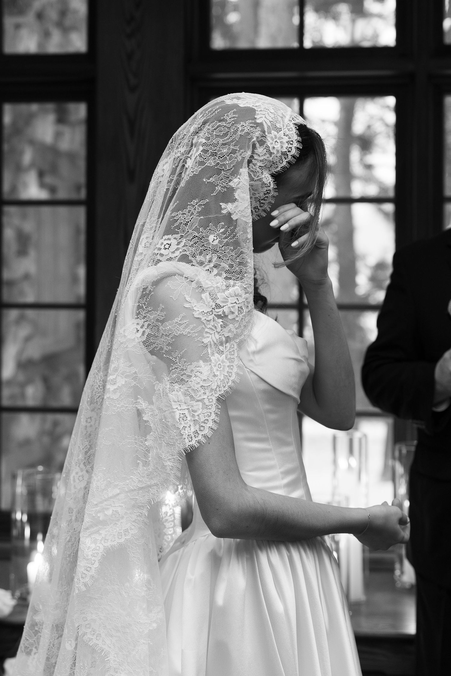 Black and white photo of an emotional bride in lace veil covering her face during wedding ceremony.