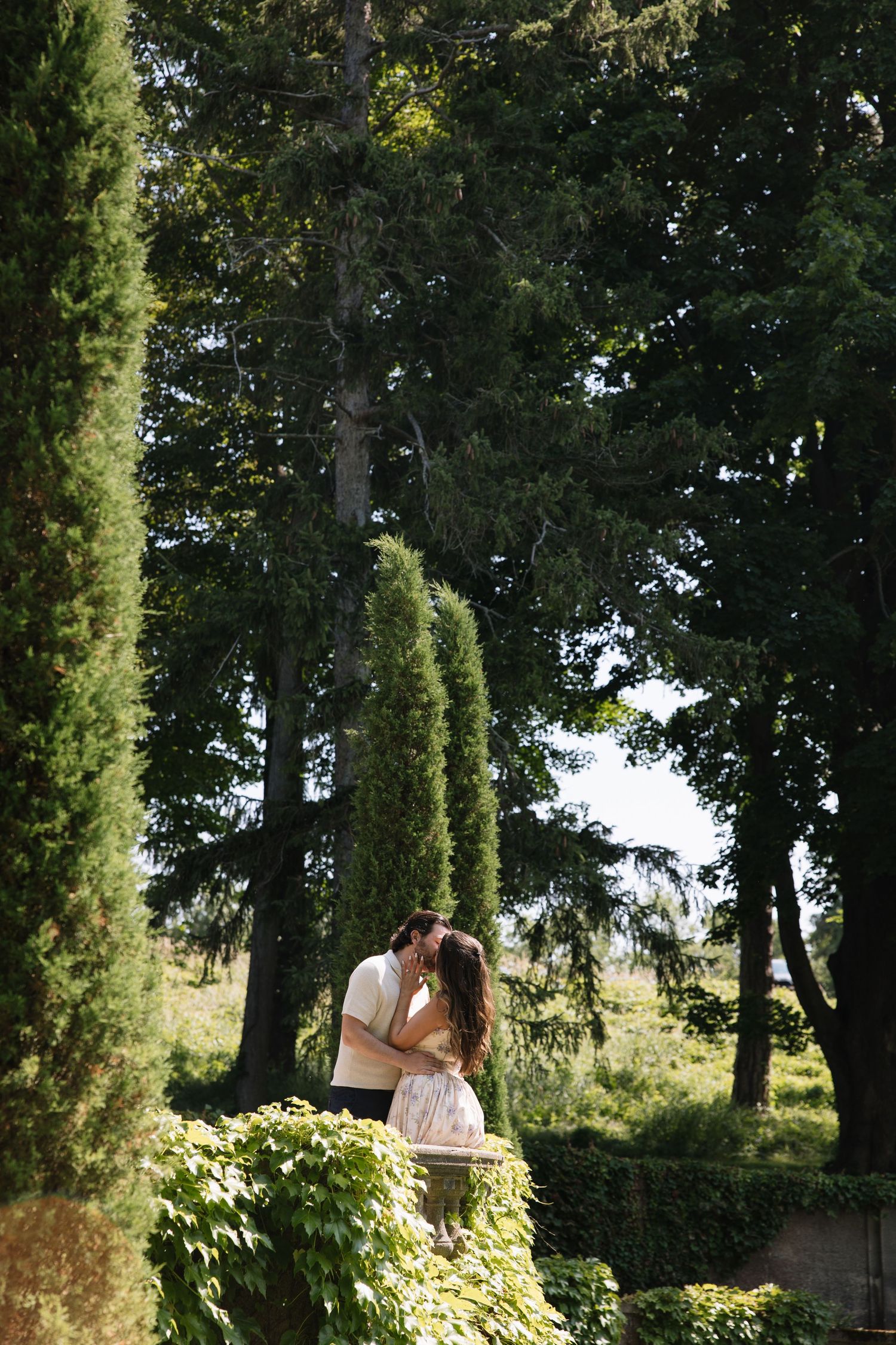 Couple sharing a romantic kiss surrounded by tall cypress and evergreen trees in a lush garden setting.