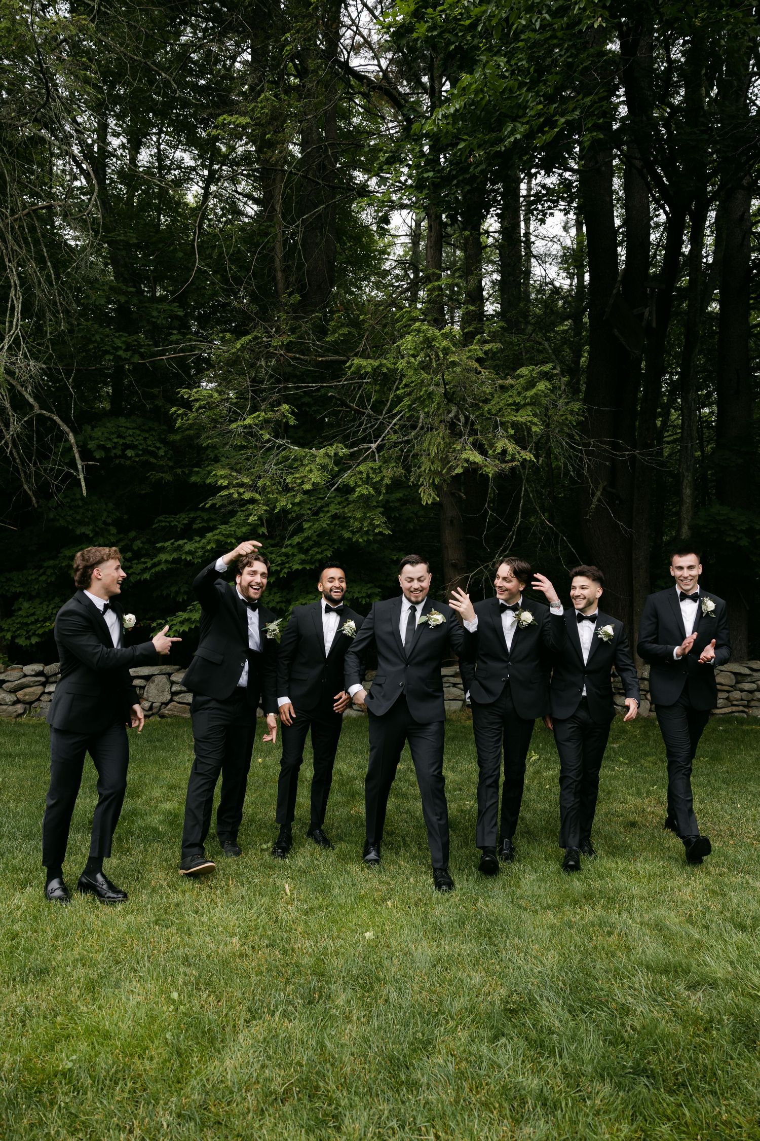 Groomsmen in black tuxedos pose in line on grassy lawn with trees in background.
