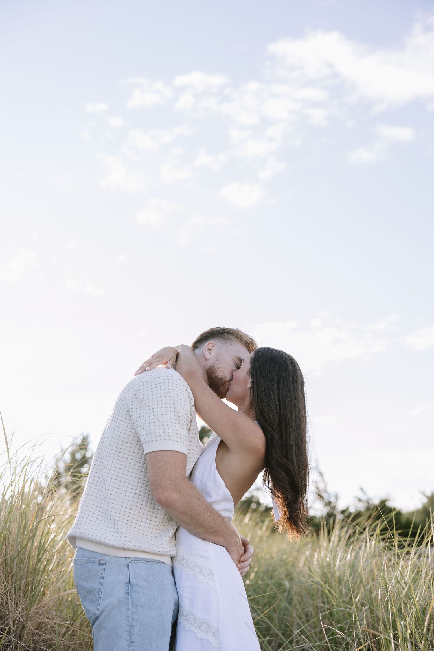 A couple sharing a romantic kiss in a field of tall grass under a bright blue sky.